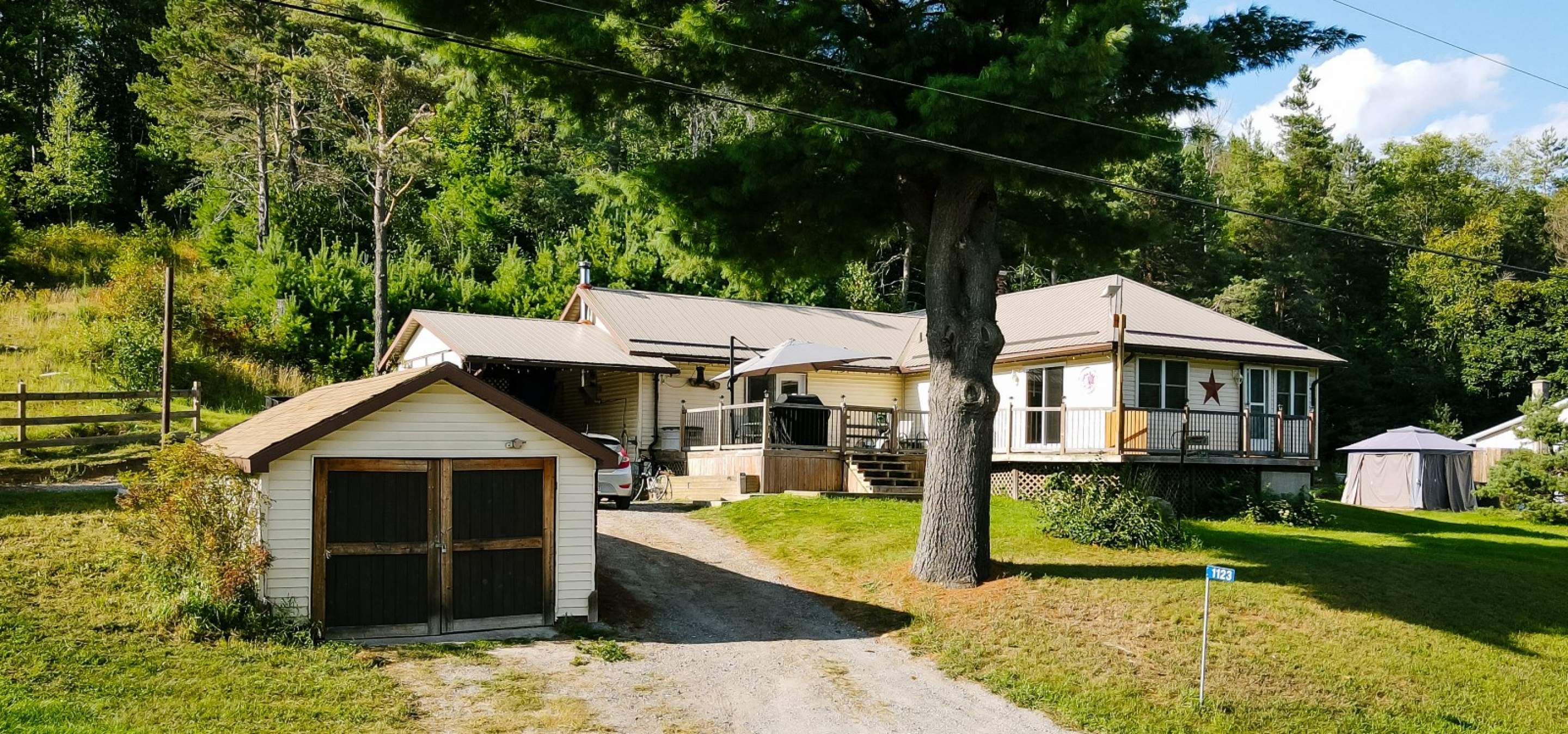 Home surrounded by trees with a grass front lawn and large deck. Gravel laneway leading to home and garage.