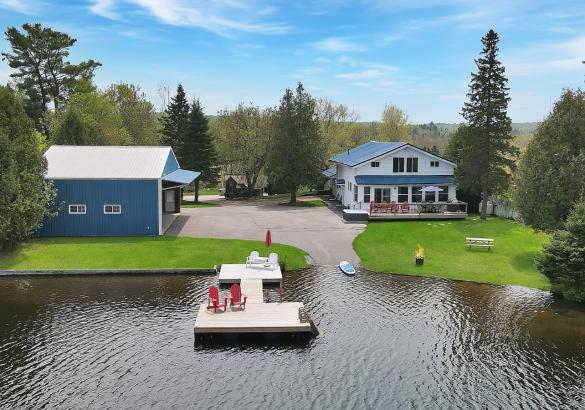 Blue airplane hanger, paved laneway, white house with blue roof. Green grass, waterfront, with a dock that has 2 red muskoka chairs.