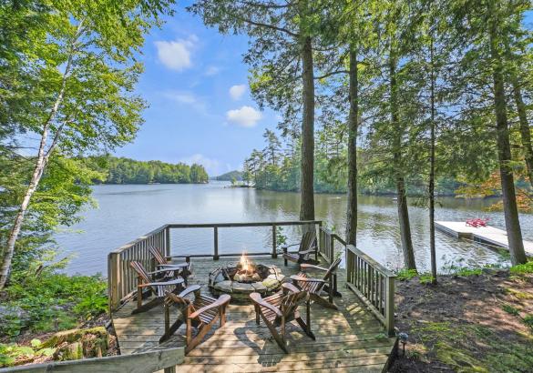 Fire pit with 6 chairs around it and a glass railing overloooking a lake. Trees surround and a dock in the water with red chairs.