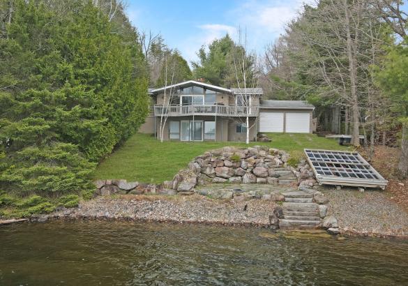 Cottage with 2 story deck, 2 white garage doors, grass hill turns into stone and a dock laying on the laneway. Trees down both sides.