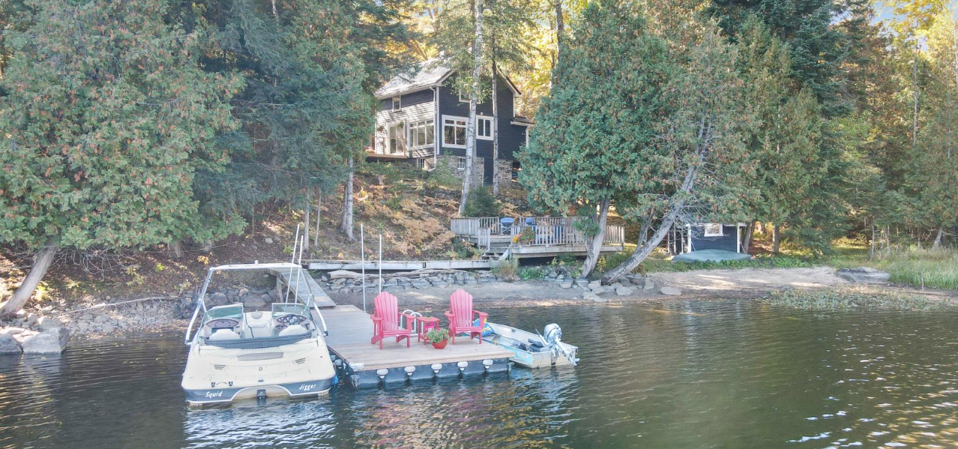 look at a cottage from the lake with 2 boats and 2 red chairs on the dock.