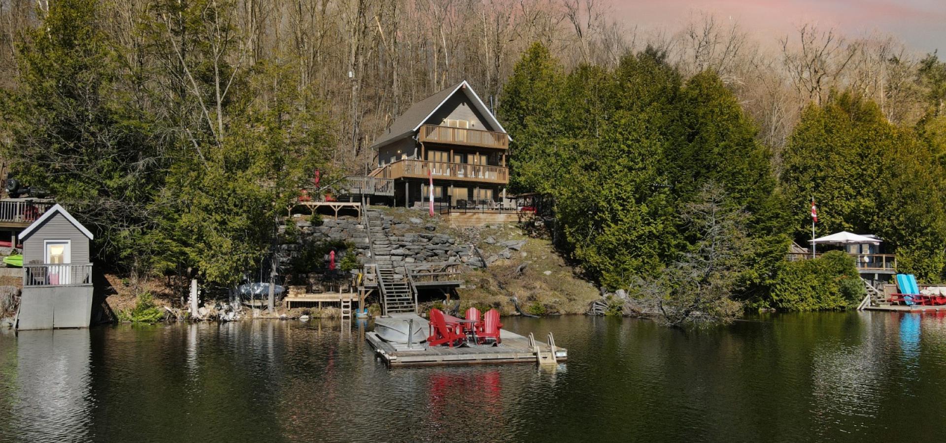 Cottage hidden amongst trees, dock out front with 4 red chairs.