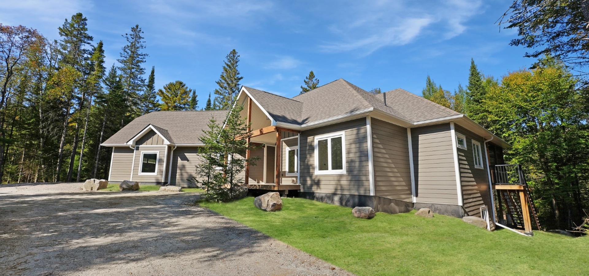 Beige siding home with white framed windows surrounded by forest, green lawn and gravel driveway in front. 
