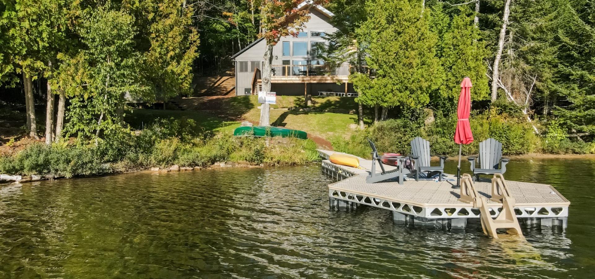 New cottage surrounded by trees with grass lot leading to the lake. Dock out front with 3 Muskoka chairs. 