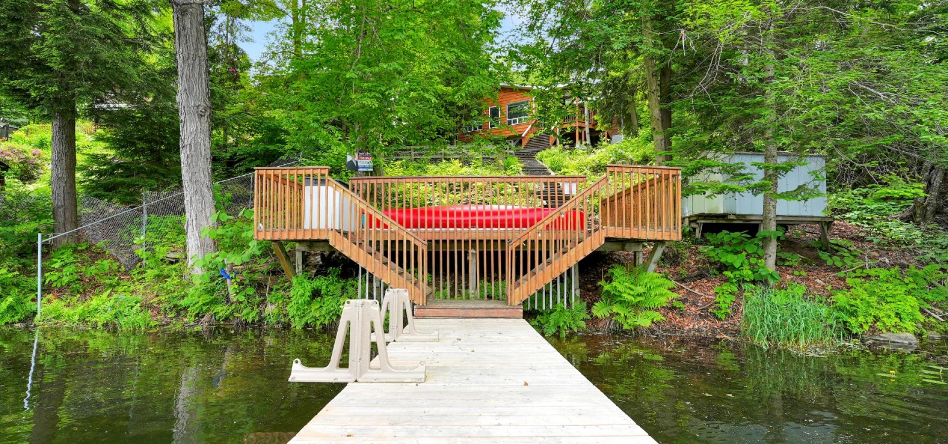 Cottage on a hill behind trees, stairs down to the dock in the water, and a red canoe.