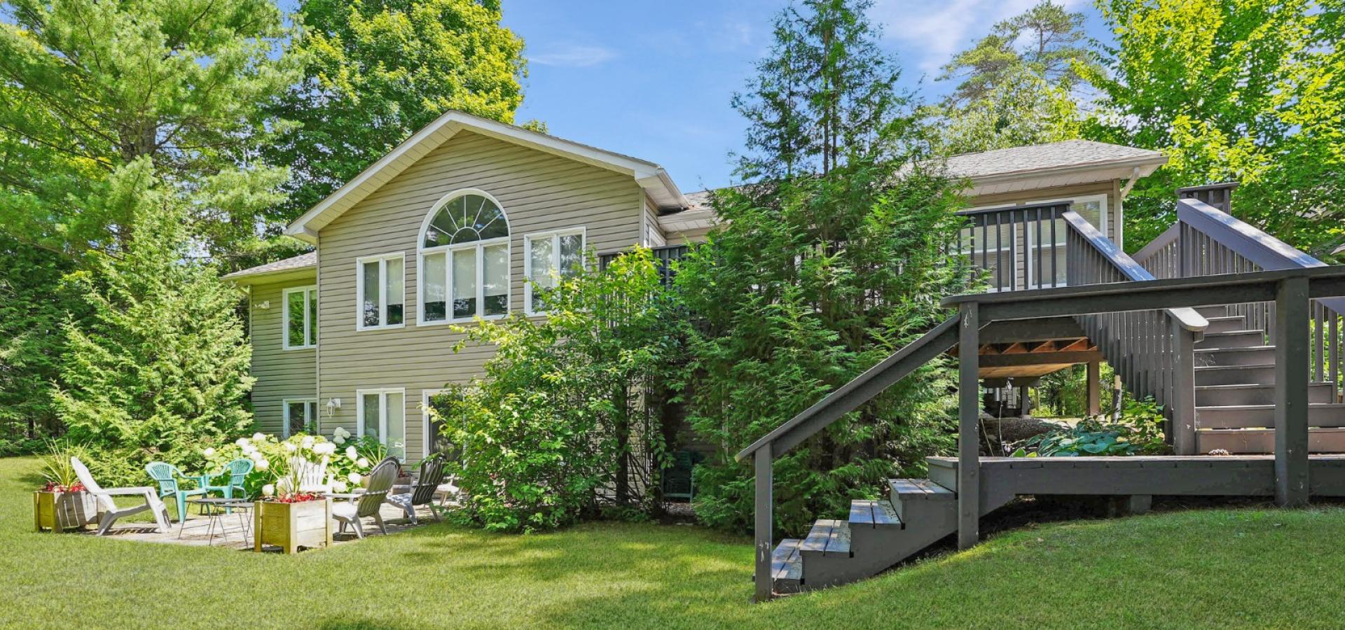 2 level home with deck and stairs on the right to green manicured lawn and patio with chairs at the front of the home