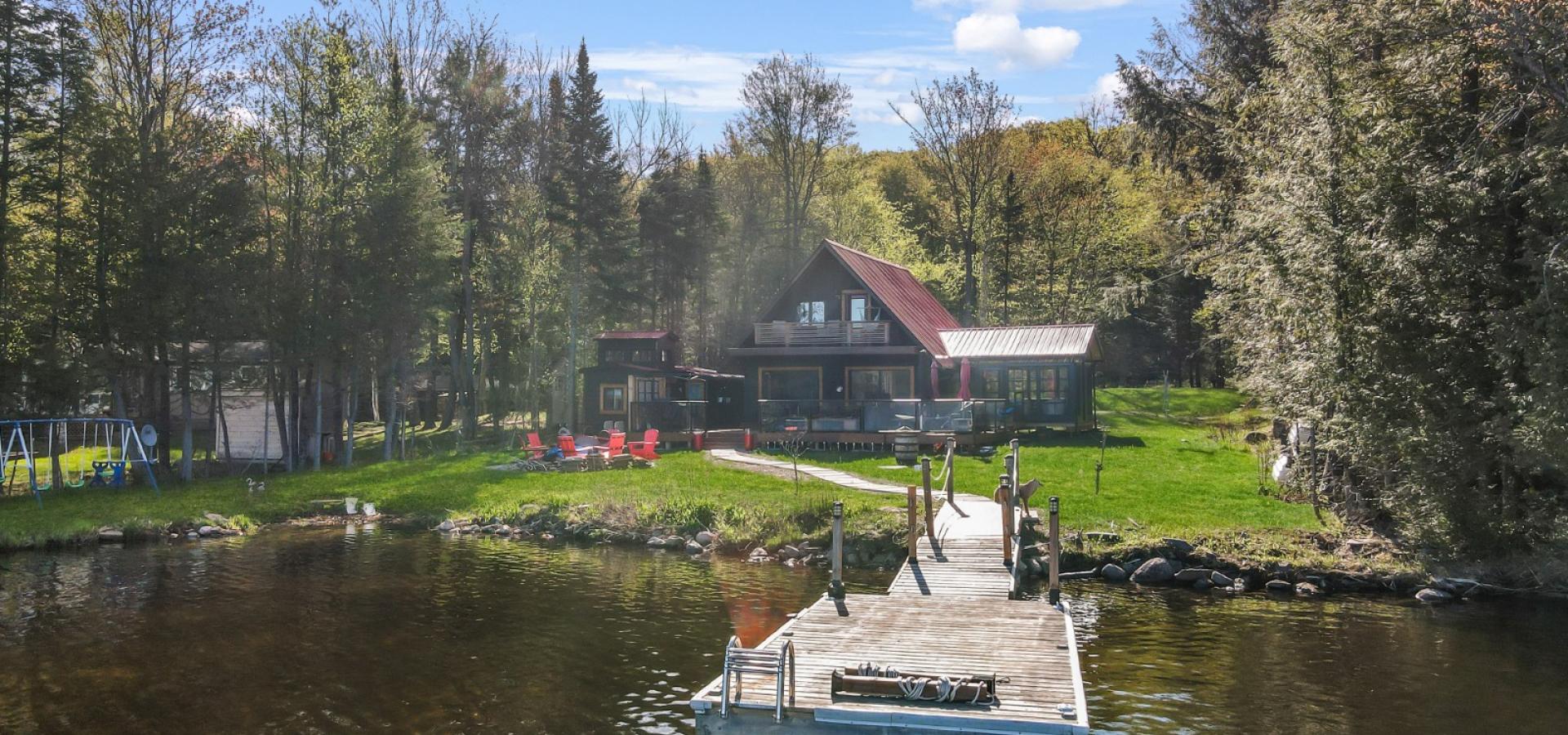 a frame cottage with green grass, red chairs around the fire, sand beach and dock into lake.