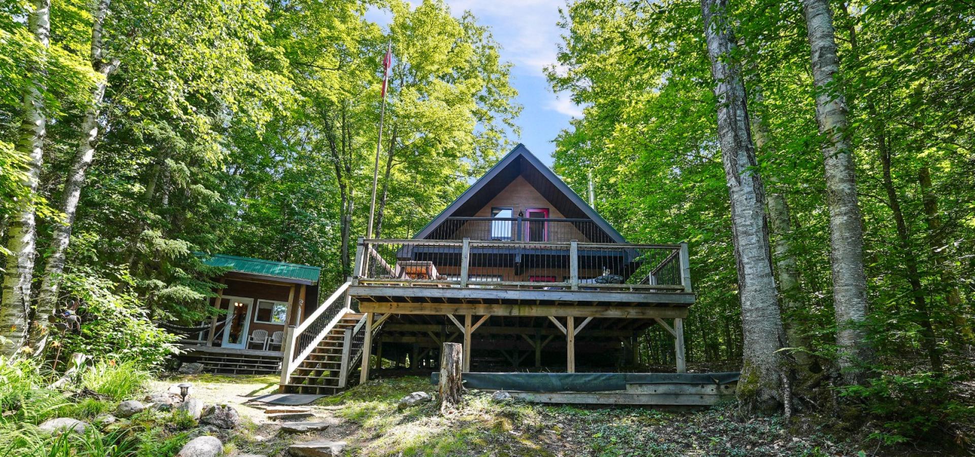 Chalet style cottage with large front deck, bunkie to the left and surrounded by trees
