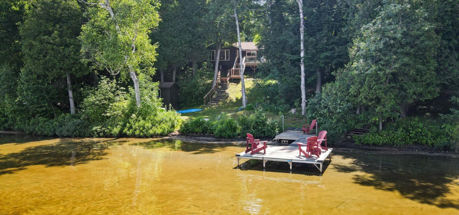 Cottage through the trees with a dock that has 4 red muskoka chairs and sandy beach