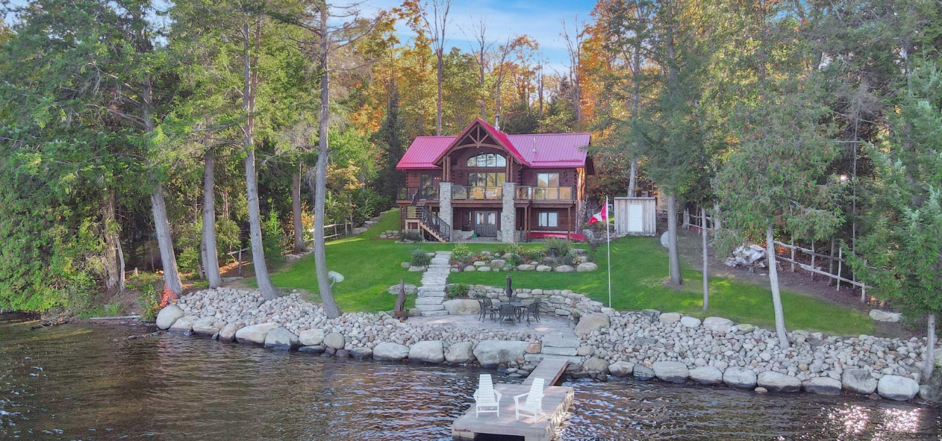 log home with red roof, green grass lawn, rock line before the lake. 