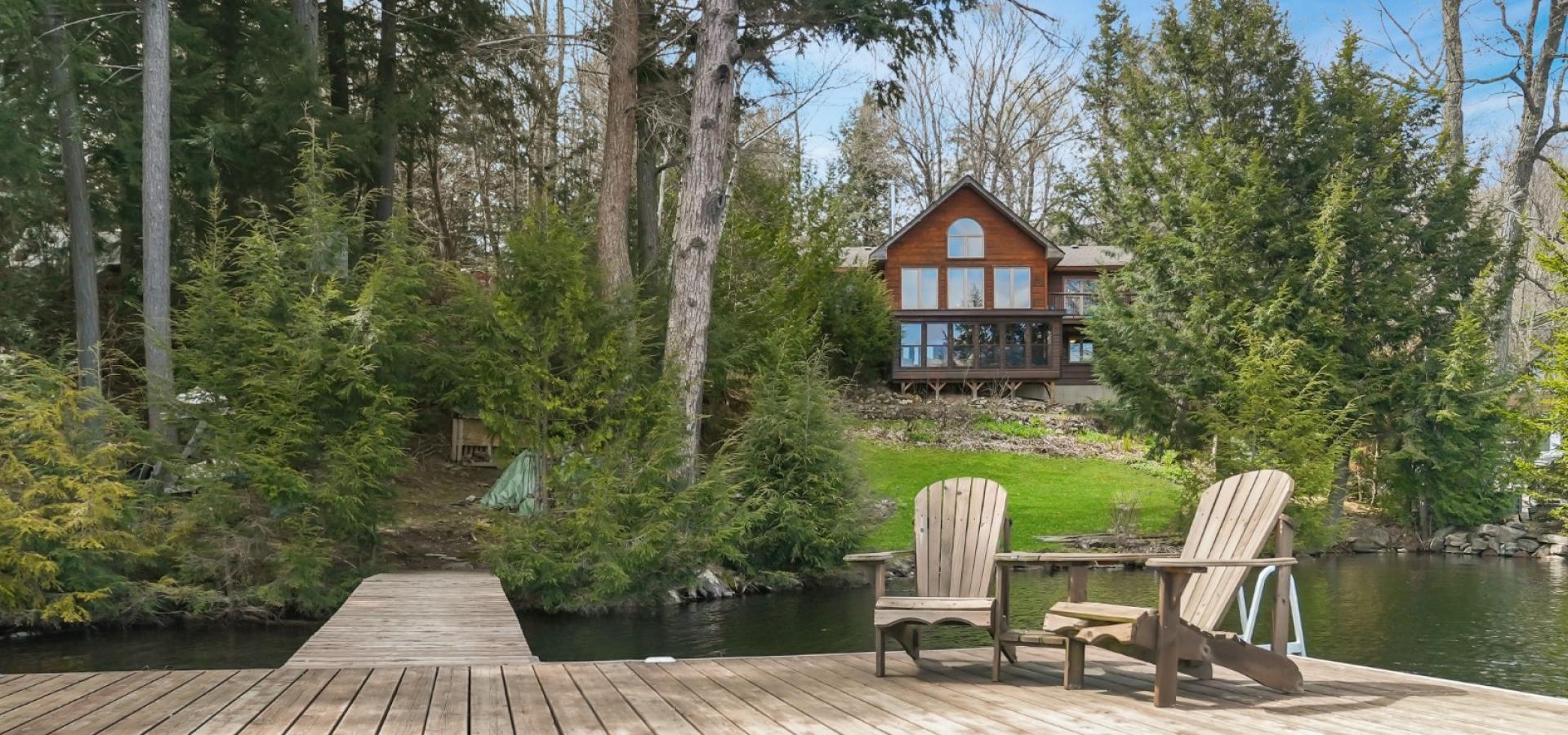 Cottage in the background with a lot of windows, surrounded by trees and a dock in front with 2 Muskoka chairs.