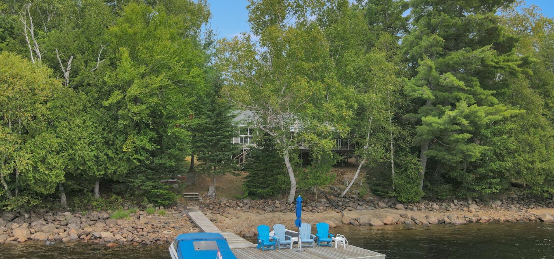 Cottage hidden behind trees with sandy beach front and dock with 4 blue chairs and boat with blue cover.