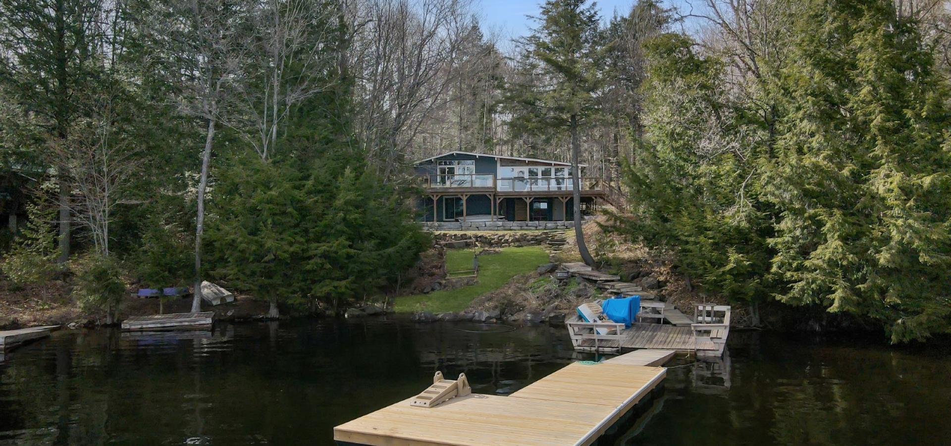 Cottage surrounded by trees, green grass in front and a dock into the water.