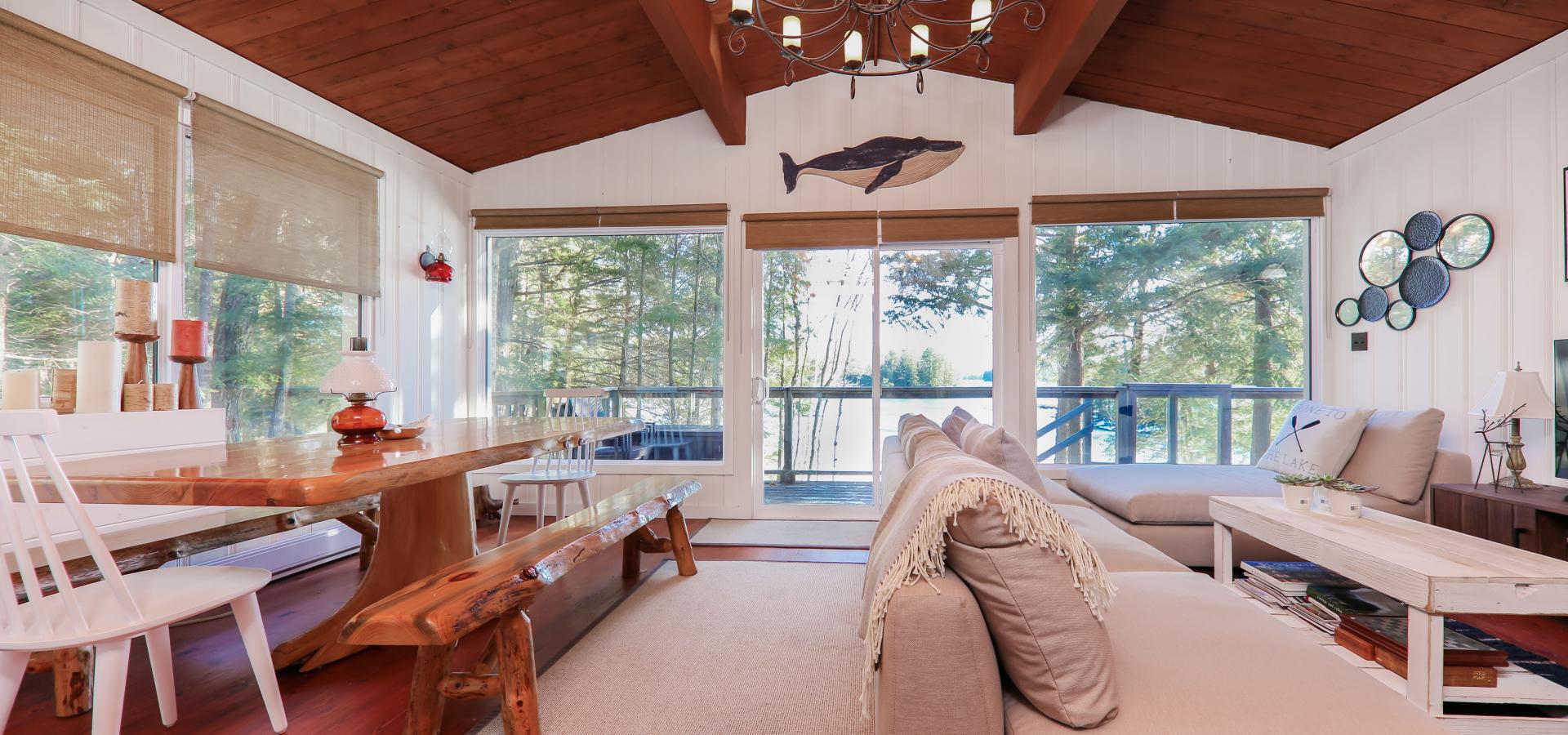 interior of a cottage with wood floors, wood ceiling, live edge dinning table with bench and 5 big windows looking at trees. 