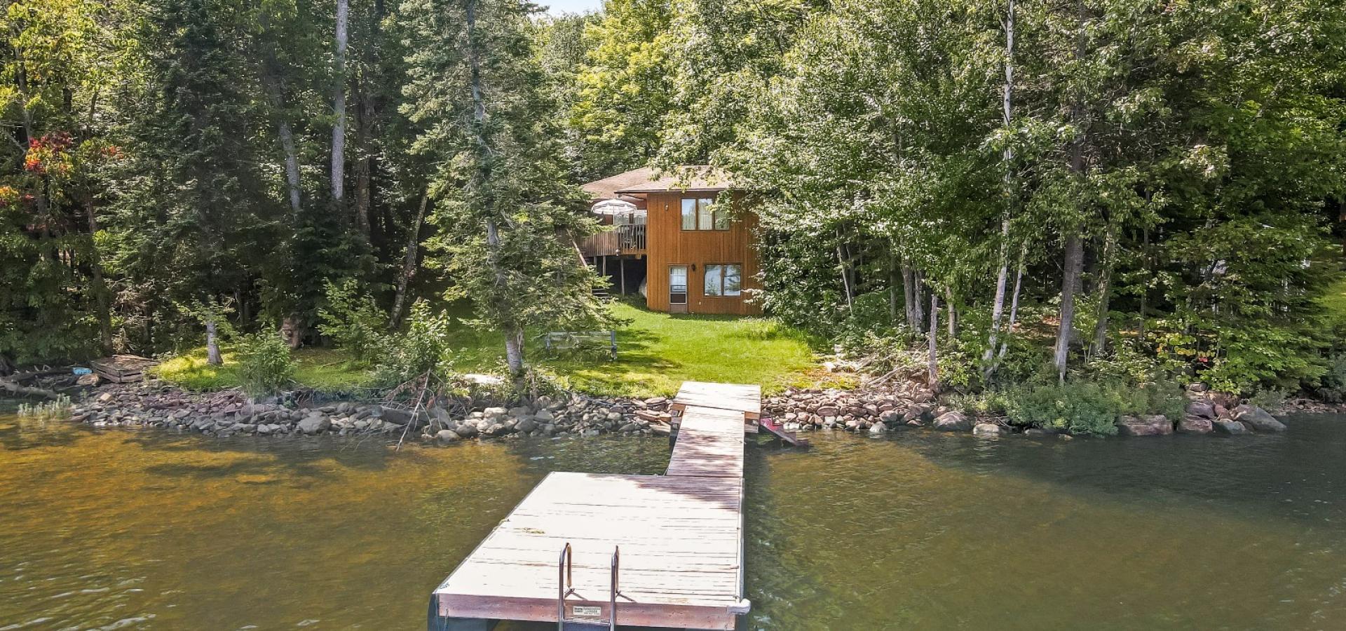 Brown wood cottage behind trees, long dock with ladder on the end