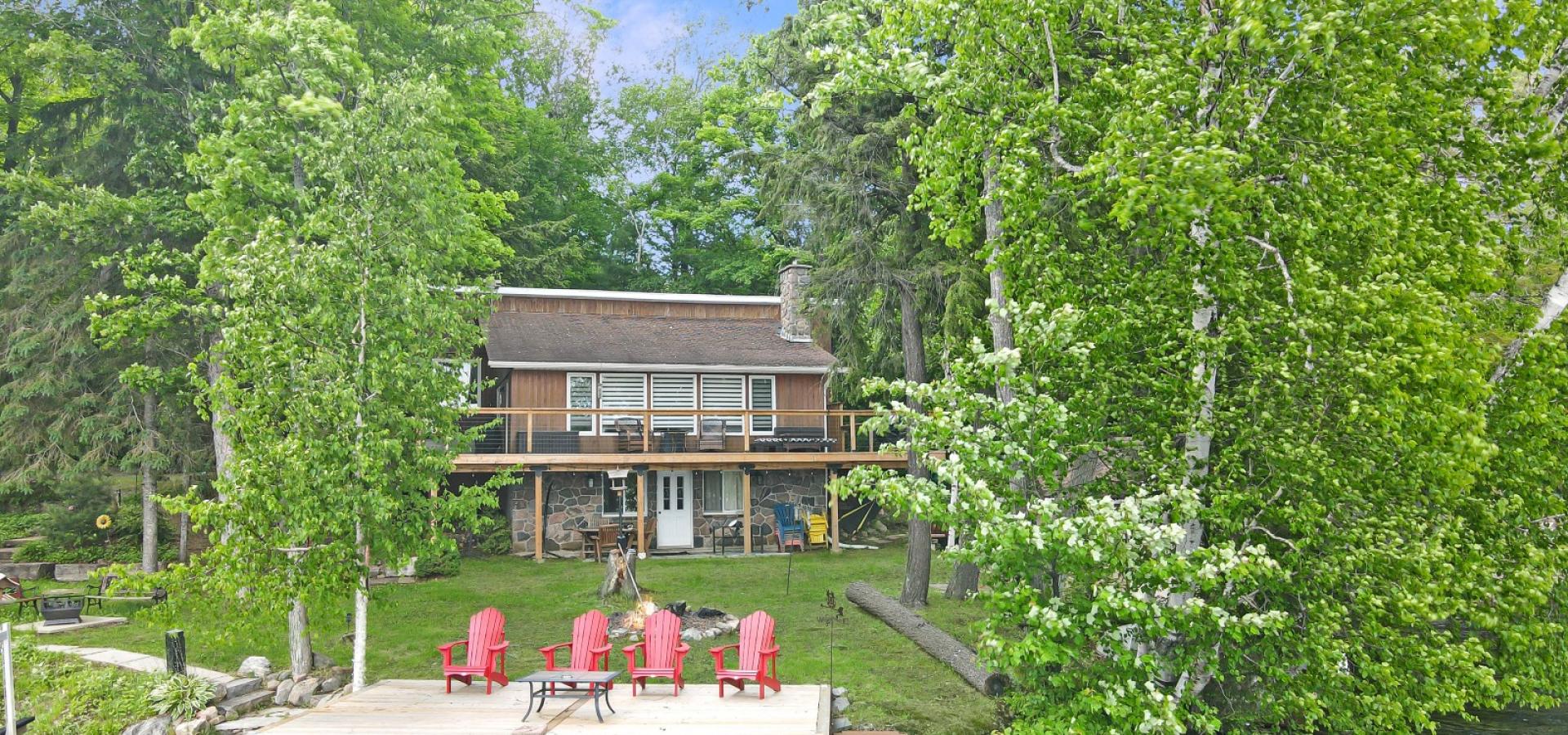 Brown cottage with 2 decks, 4 red muskoka chairs, and large trees.