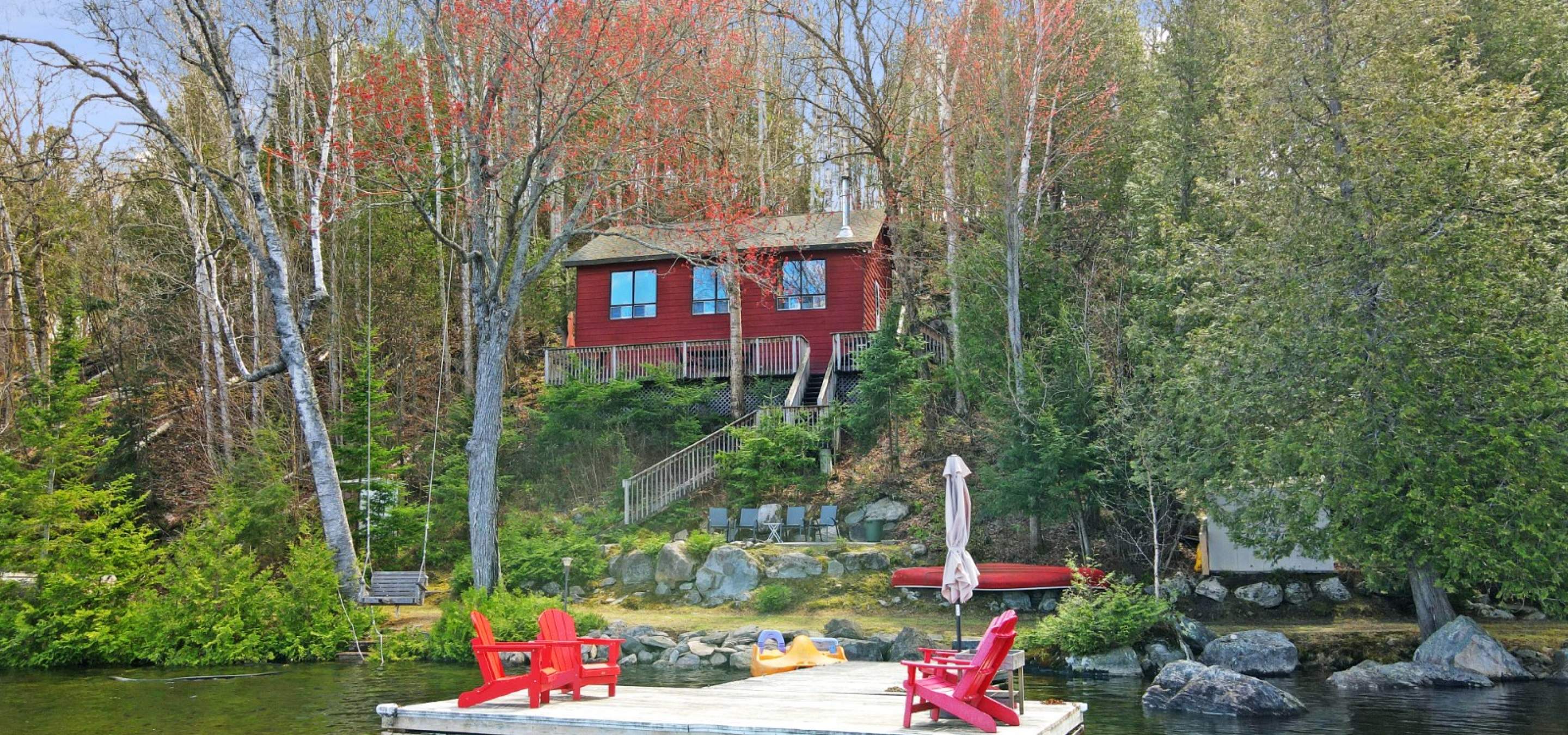 Red cottage with 3 windows, surrounded by trees. Dock in front with 4 red chairs
