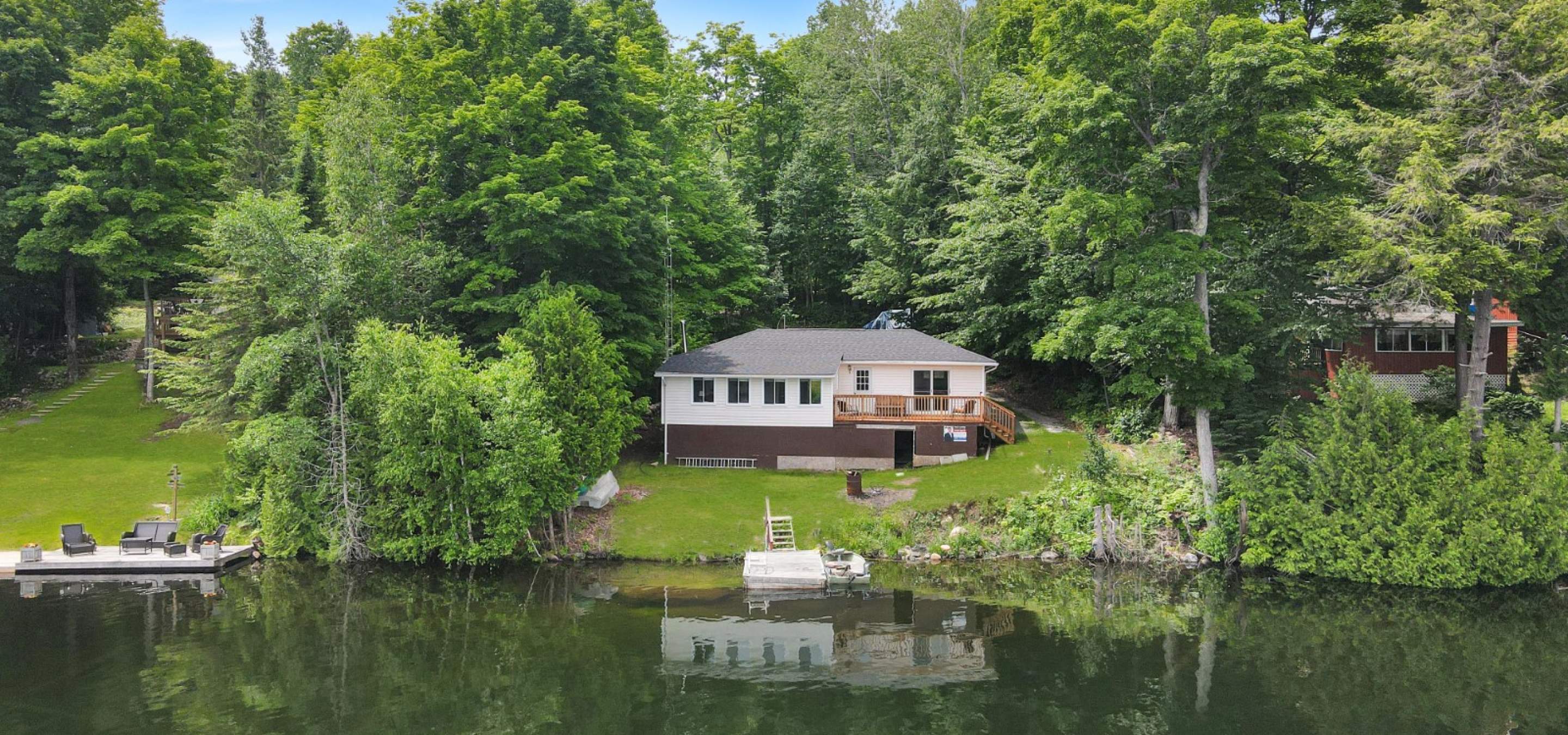 white cottage with a deck surrounded by trees and grass leading to the lake.