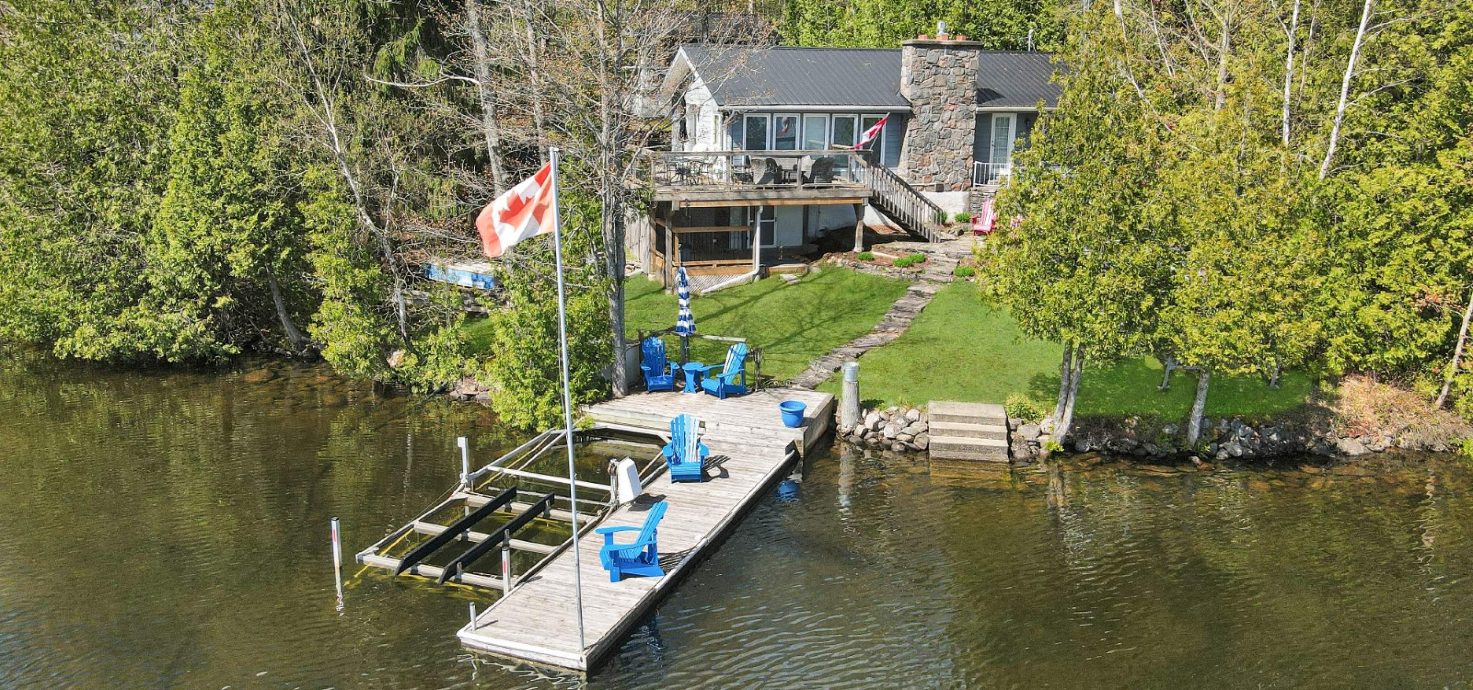 White cottage with a stone chimney with wrap around deck. Surrounded by trees with a dock in front featuring 4 blue chairs. 