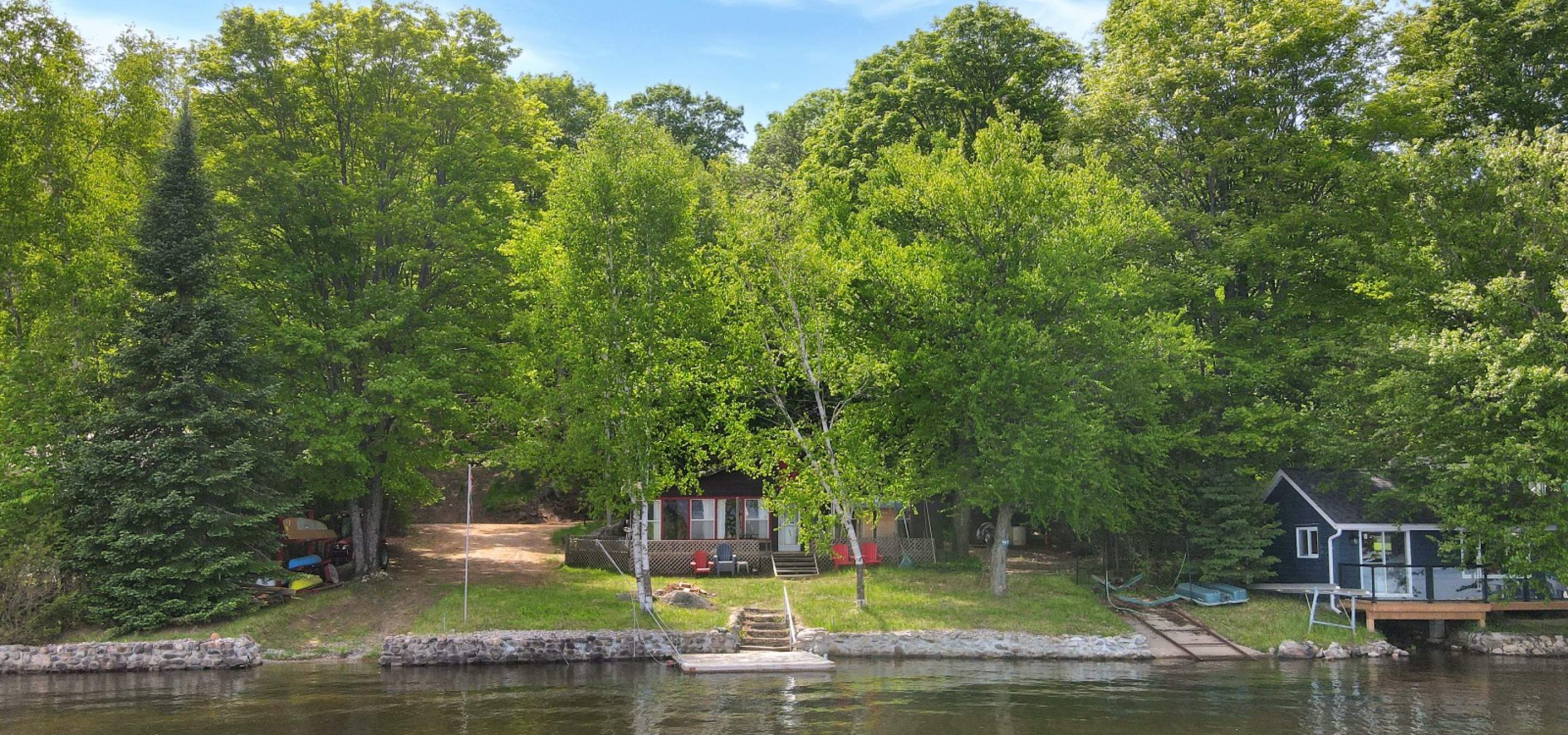 looking at a cottage from the water, large trees, stone wall, and boat launch