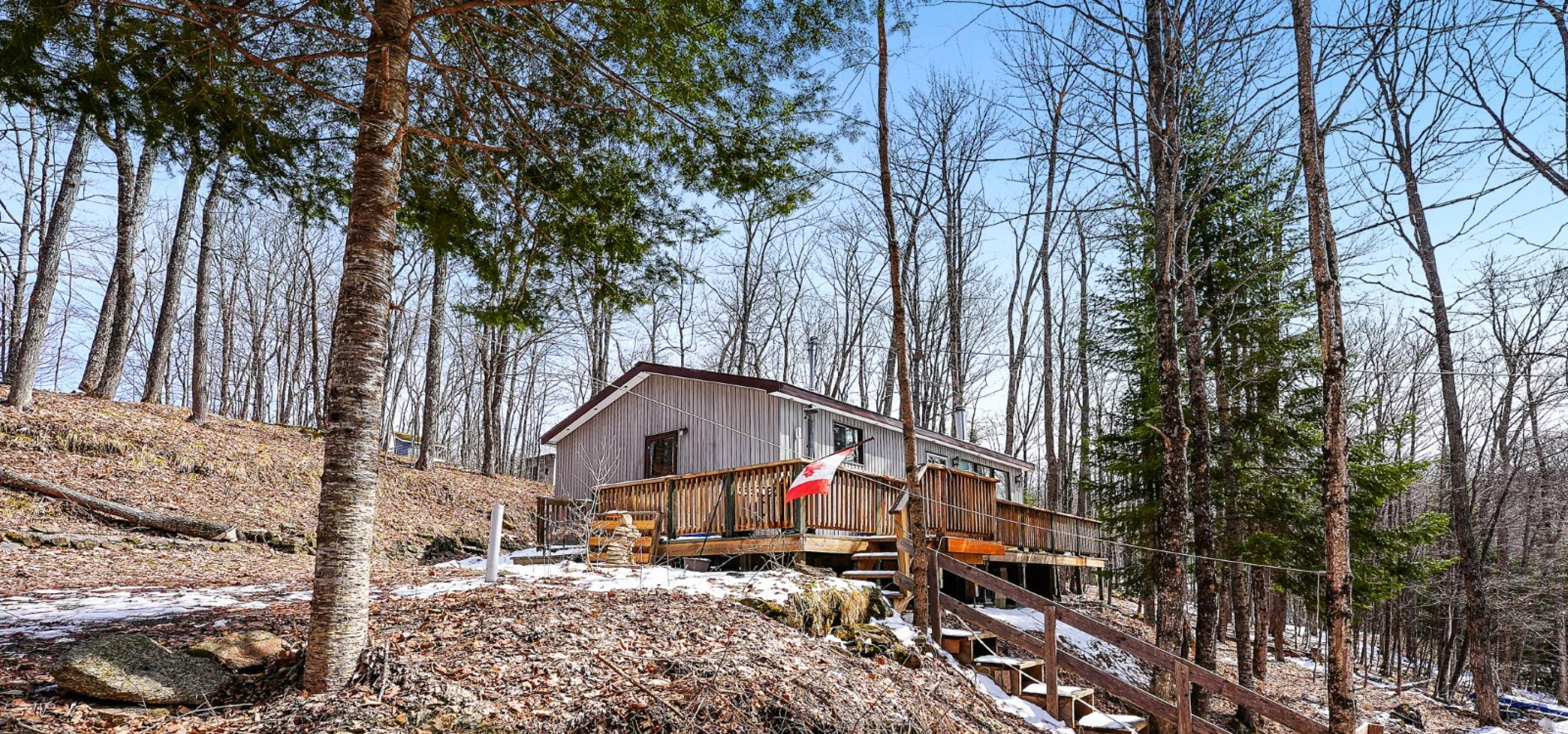 Light coloured cottage with front deck, and stairs going up the hill to it. Leaves on the ground.