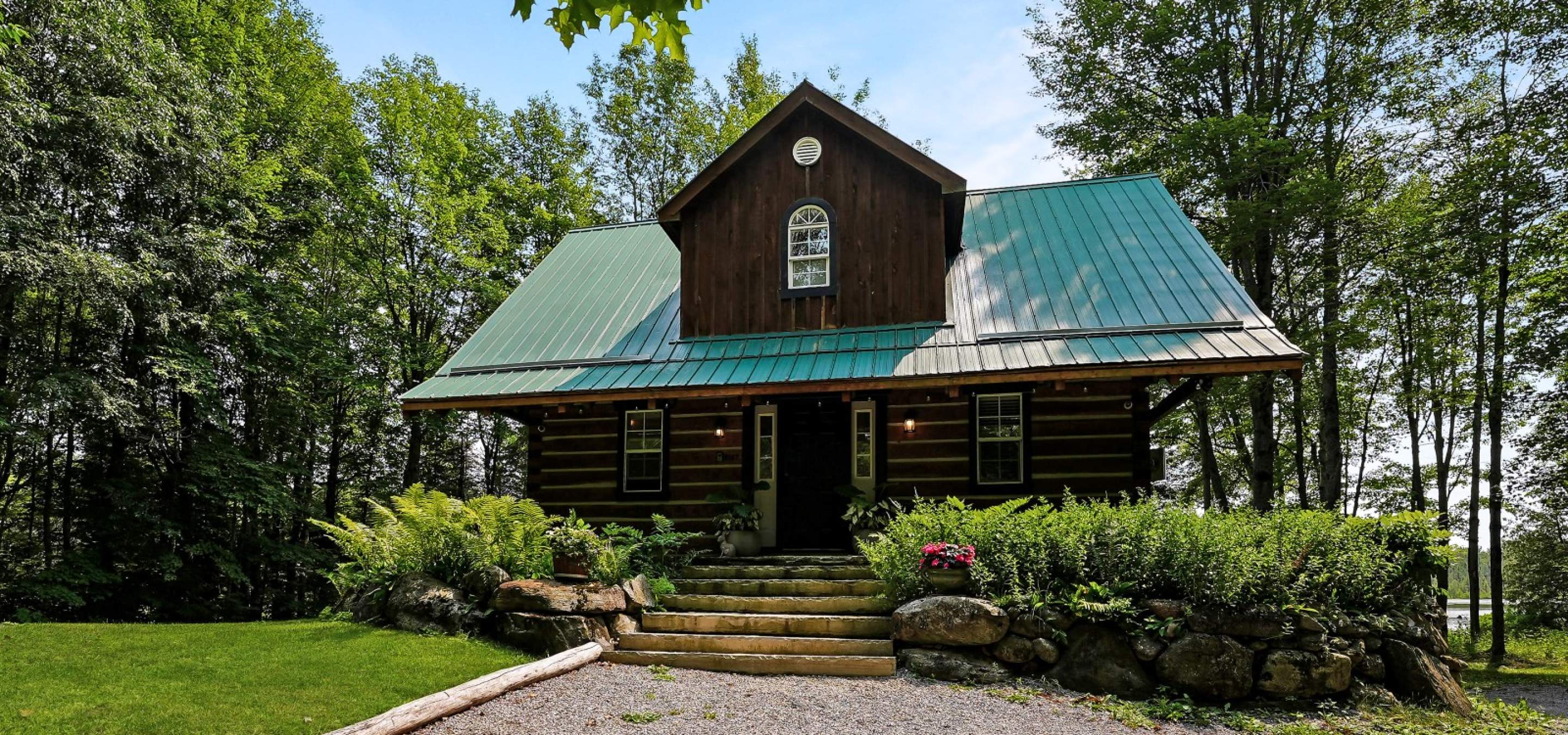 Lot home with green metal roof, stone stairs leading up too, bushes and gravel laneway