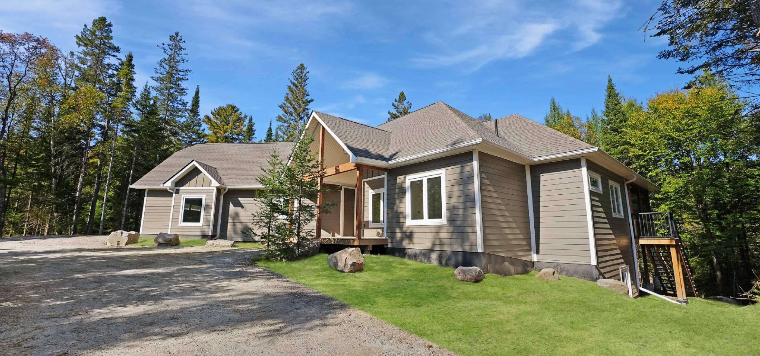 Beige siding home with white framed windows surrounded by forest, green lawn and gravel driveway in front. 