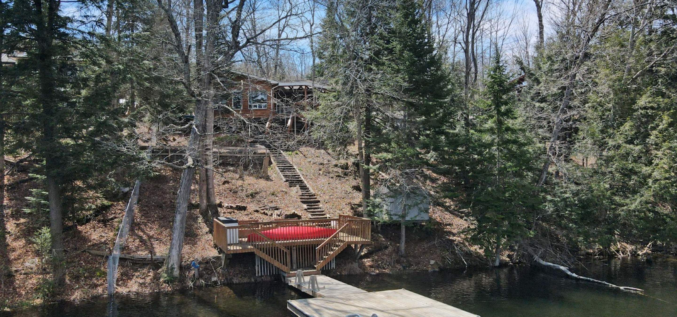 Cottage on a hill behind trees, stairs down to the dock in the water, and a red canoe.