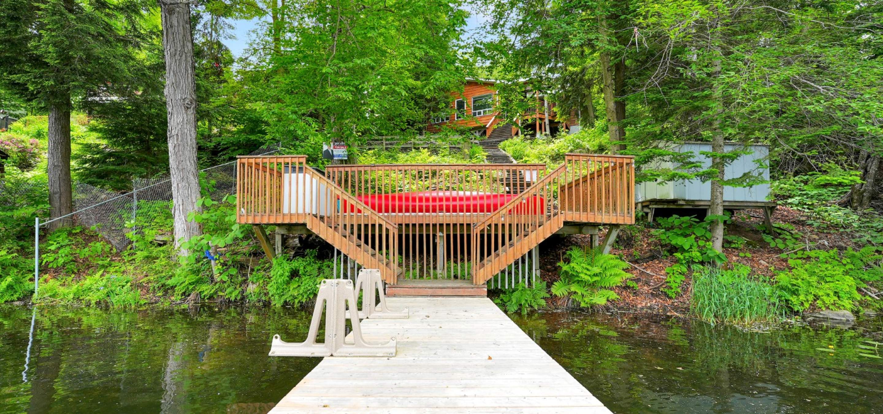 Cottage on a hill behind trees, stairs down to the dock in the water, and a red canoe.