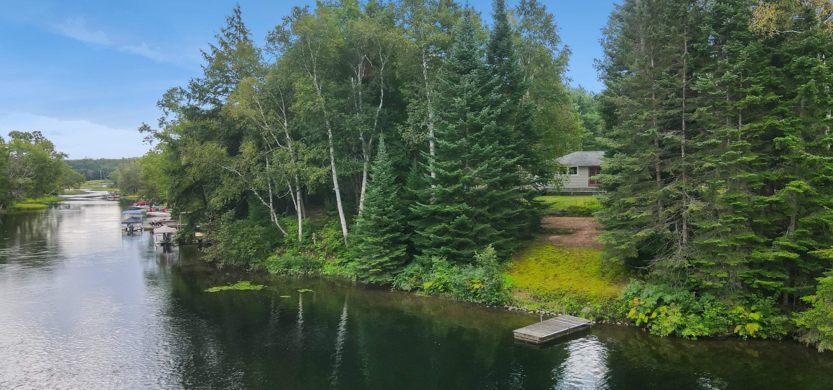 view of a grey house from the river with a dock, sandy area and trees
