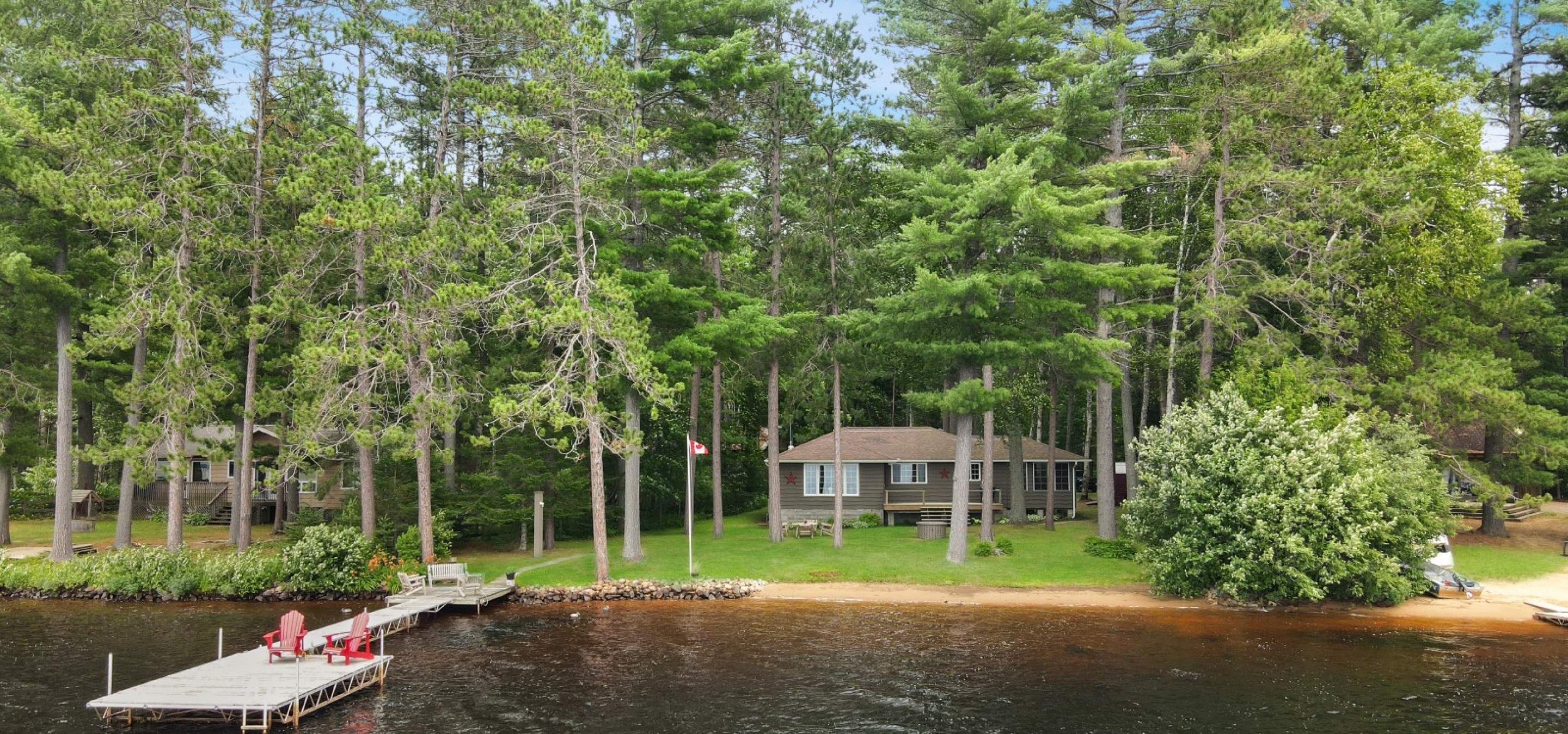 Grey cottage behind trees, sand beach and long dock with 2 red chairs