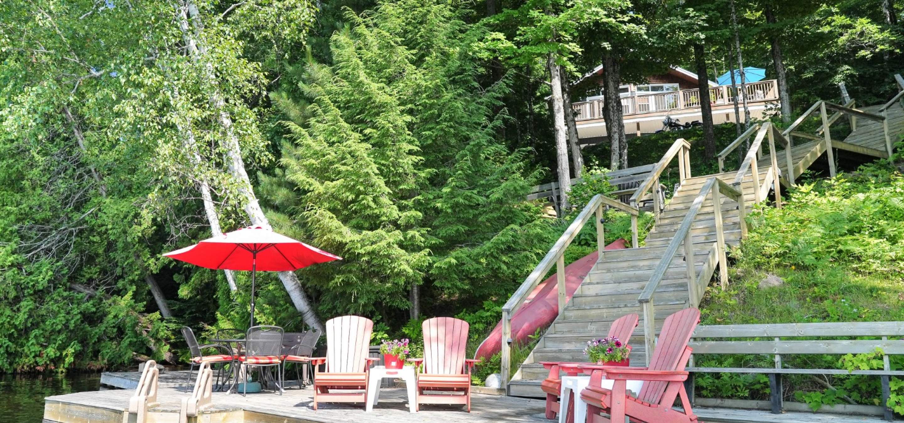 Dock with muskoka chairs with a red umbrella. Stairs leading up to the lakeside home amoungst trees