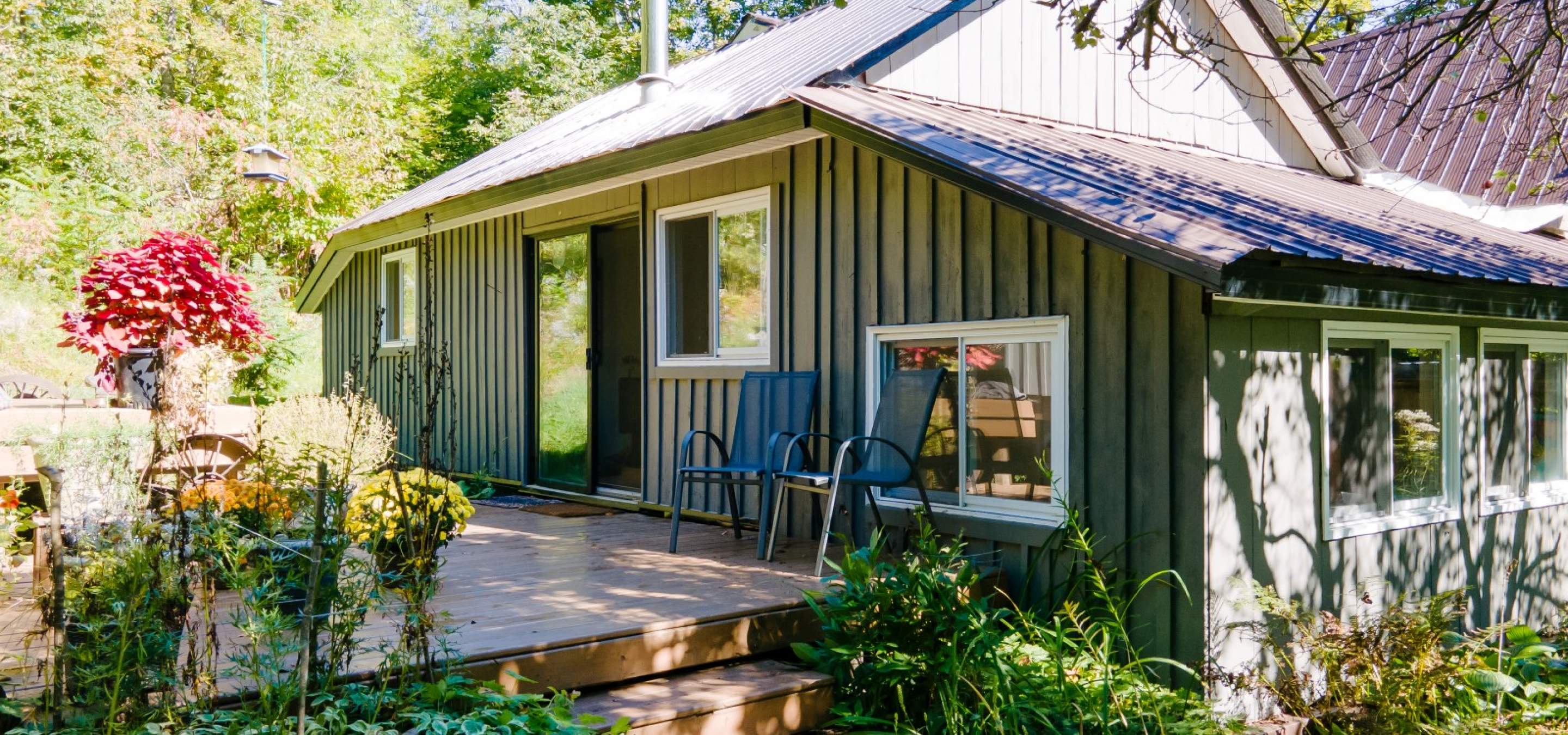 Home with a deck and 2 chairs and surrounded by gardens and trees.