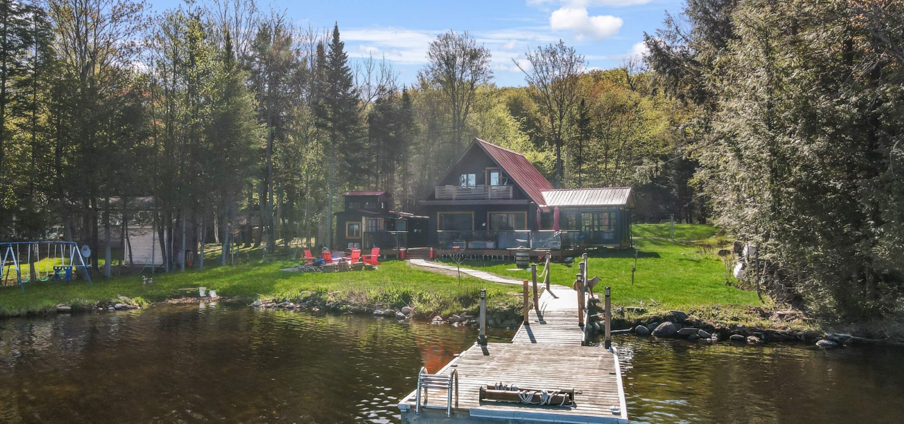 a frame cottage with green grass, red chairs around the fire, sand beach and dock into lake.