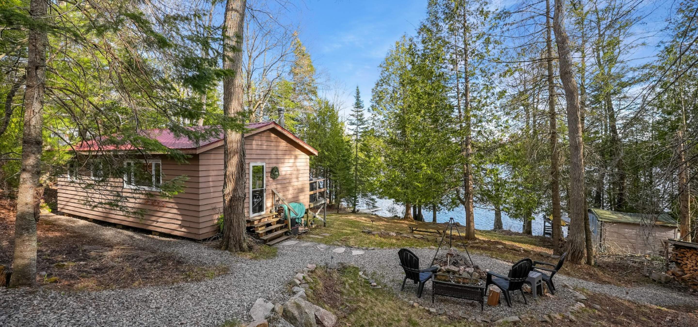 View of the back of a cottage with stone path leading to chairs around a fire and lake in the back