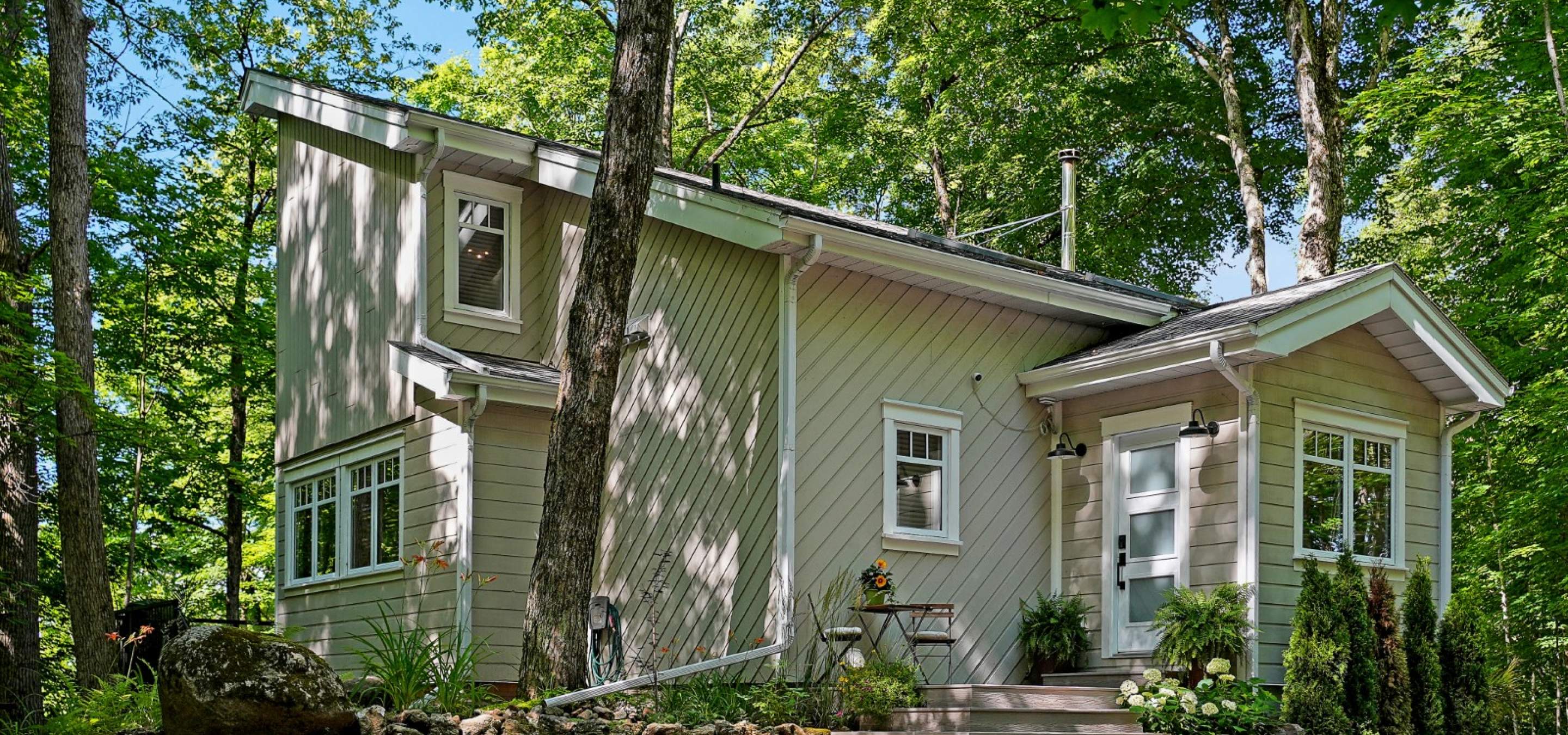 Cottage surrounded by mature trees, featuring a deck with bistro table set and landscaped gardens.