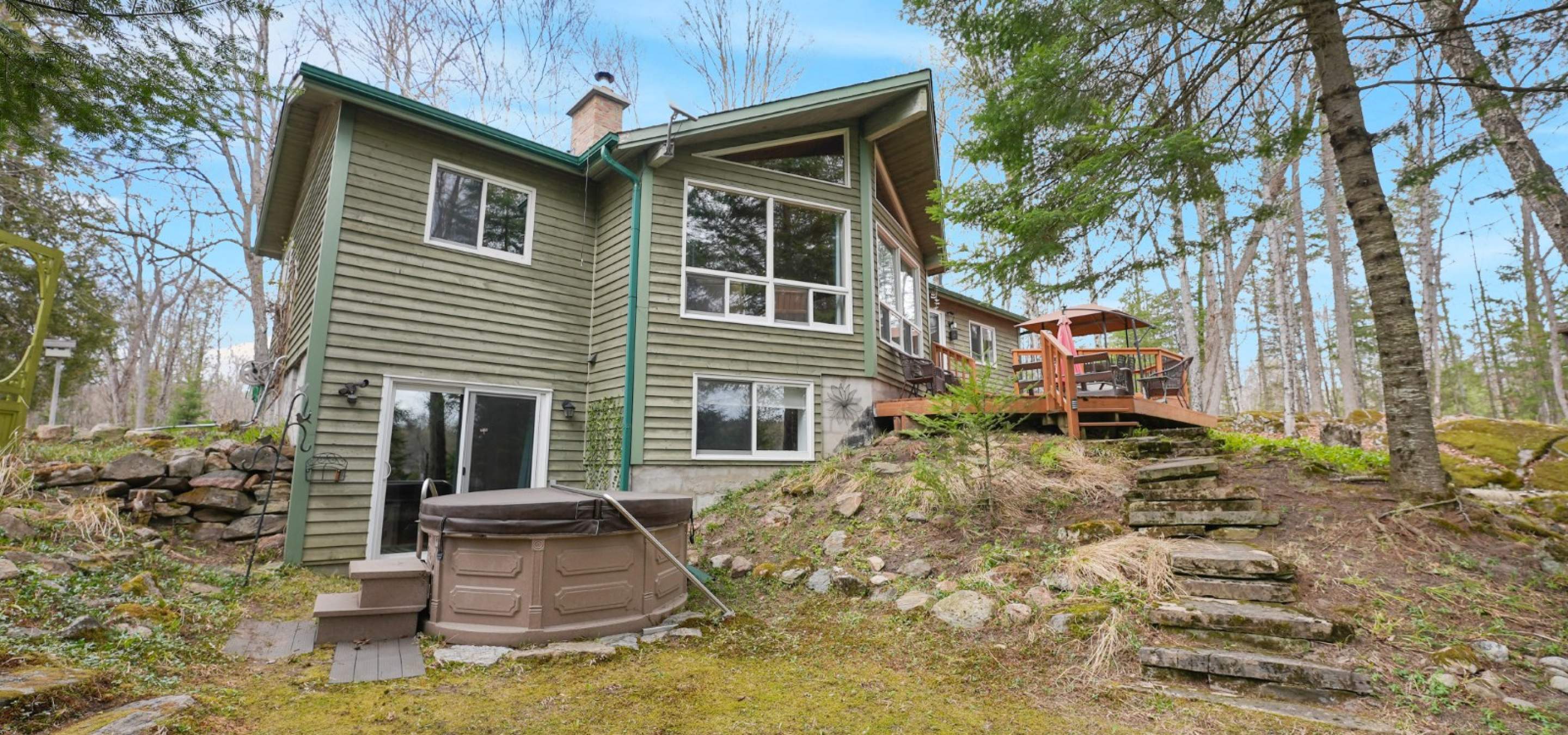 green cottage with walkout basement and hot tub. Stairs leading off the deck. Blue skies.