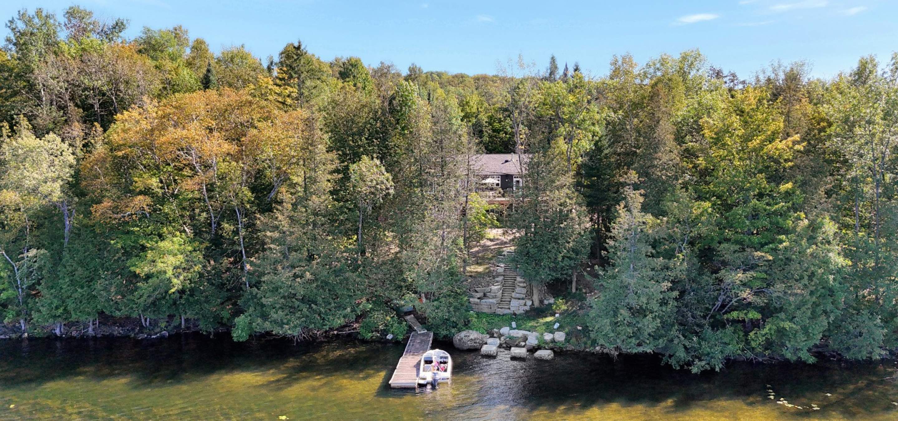 view from the lake looking at a cottage hidden behind trees, stone stairs up to it, and a dock in the lake