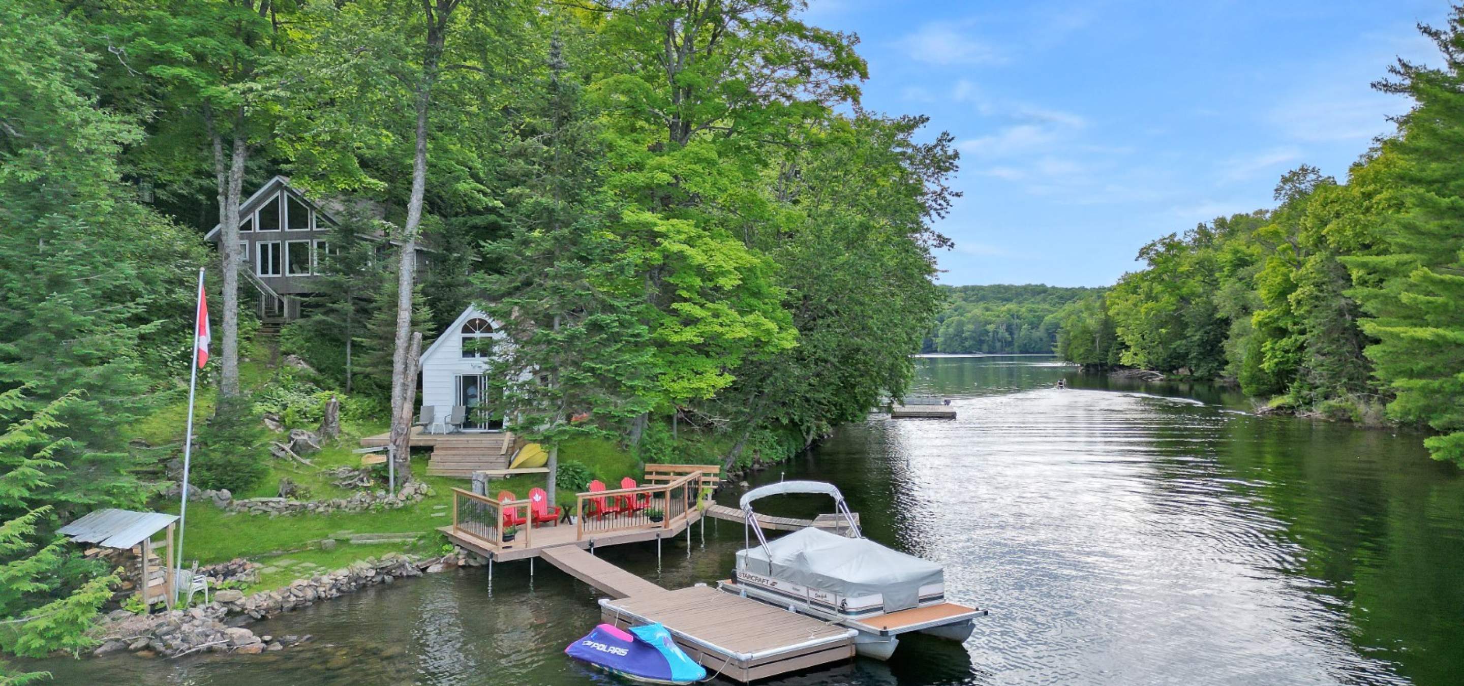 Dock with parked pontoon and jet-ski, ramp to waterside deck with muskoka chairs, a white frame bunkie and grey cottage on the hillside.