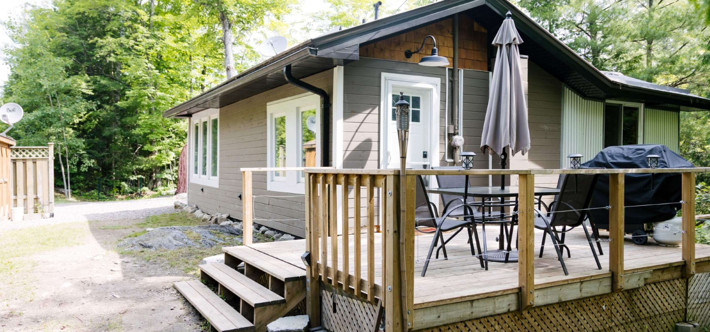 Wood deck with patio set and barbecue, tidy cottage with trees in the background.