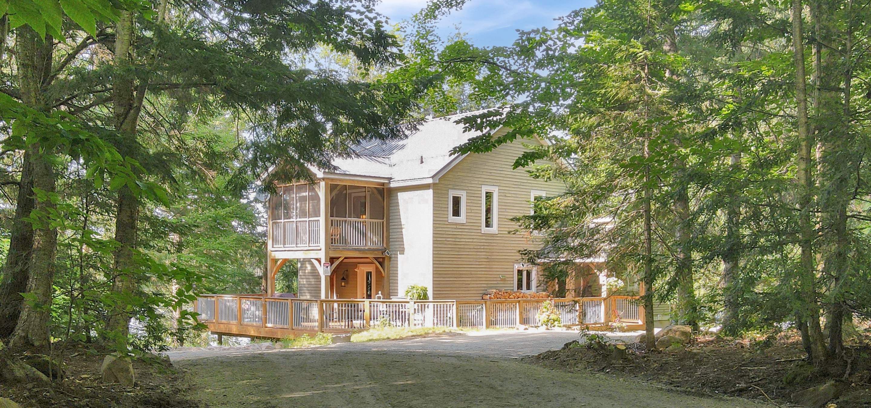 Driveway approach to this home or cottage through the trees with sun shining on the siding clad building with decking and porch