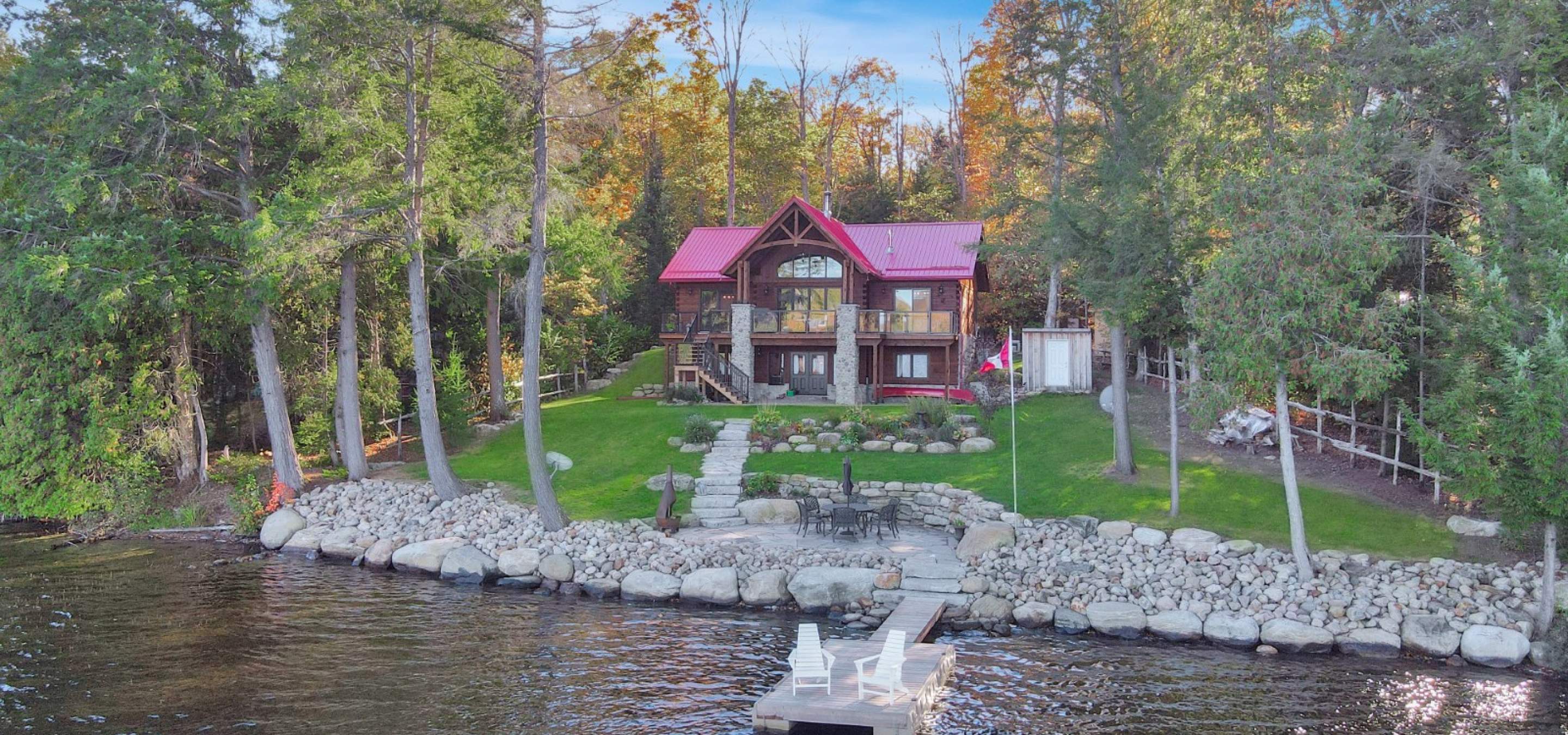 log home with red roof, green grass lawn, rock line before the lake. 