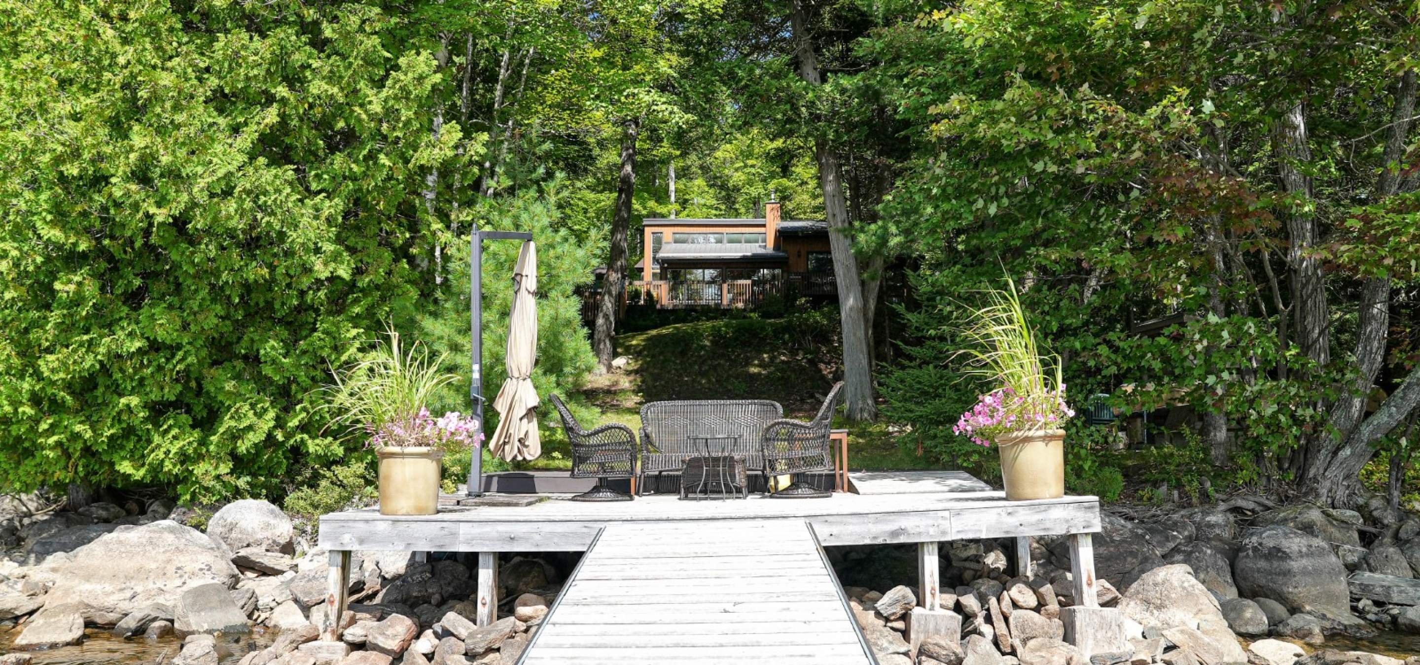 Cottage hidden amoungst the trees with a dock into the water. Rocky shoreline and seating area on the dock. 