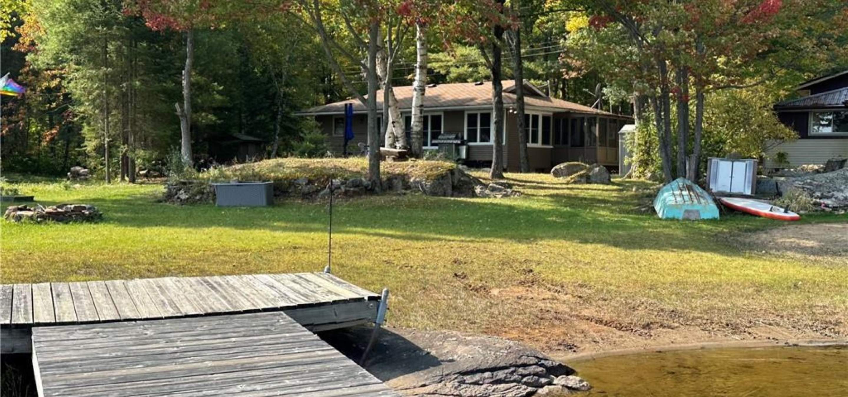 Cottage in the background with trees in front, grass and dock into the lake. Showing sandy shore as well. 