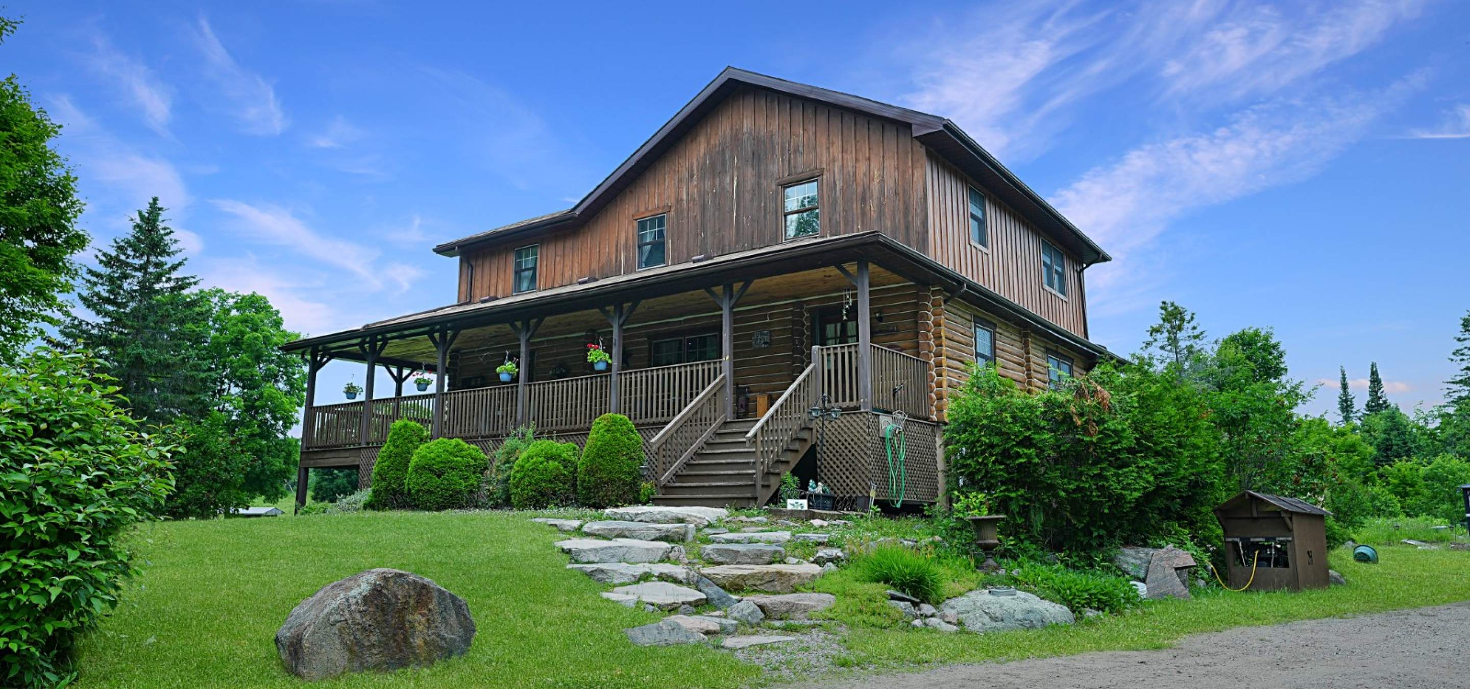 Log home with covered front porch. Stone stairs leading up to the home, surrounded by grass and trees, gravel laneway.