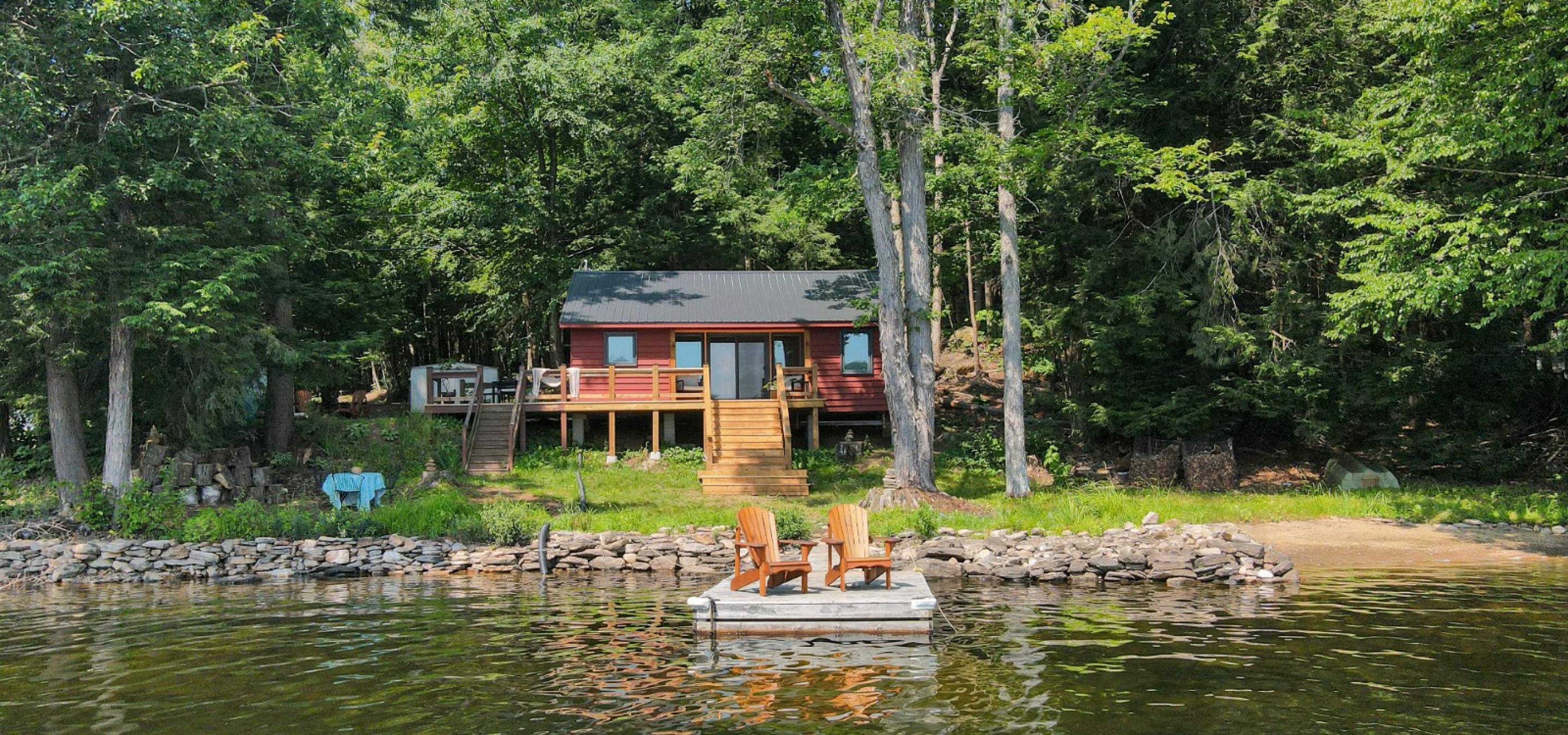 Red cottage in the trees with stairs leading down to grass. 2 muskoka chairs on dock in front.