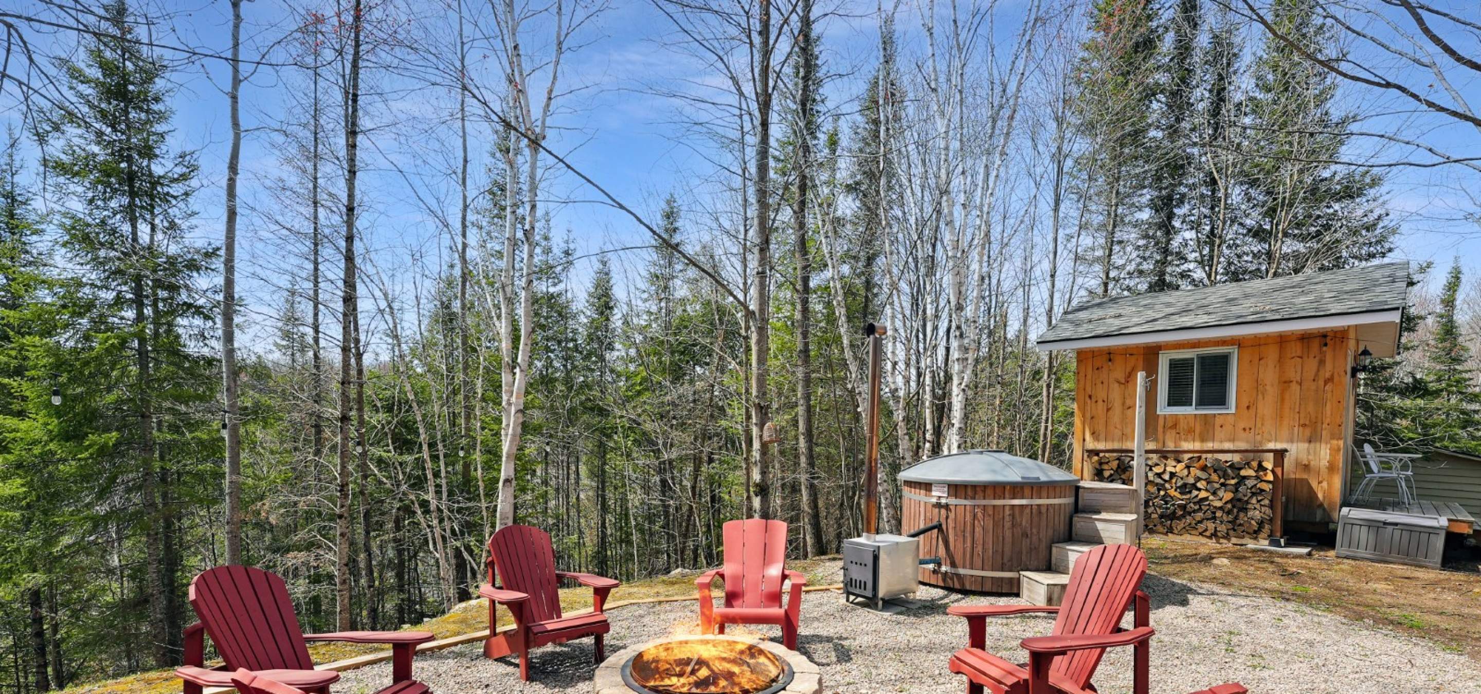 6 red muskoka chairs around a campfire and a bunkie in the background