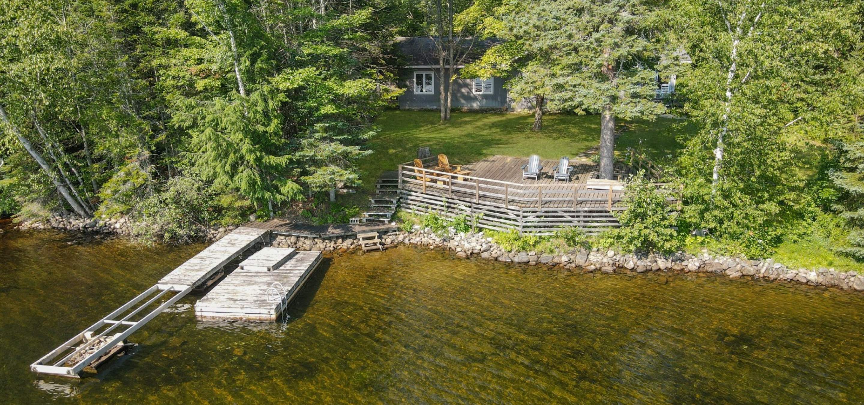 grey cottage hidden behind trees, green grass, large lake side deck and dock in the lake. 