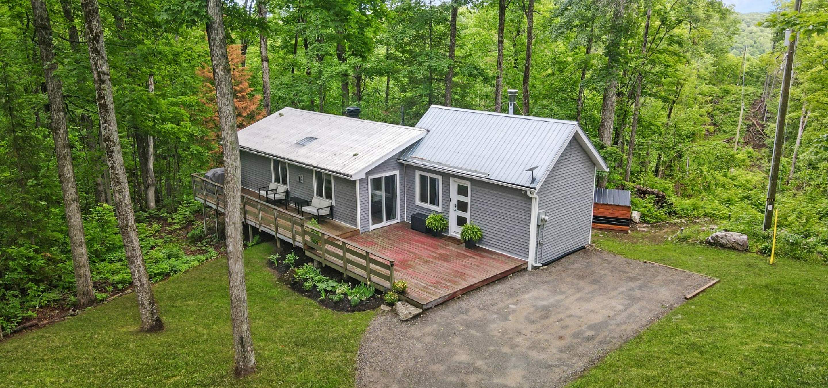 Grey home with white roof, doors and windows. Large front deck, gravel laneway and grass.