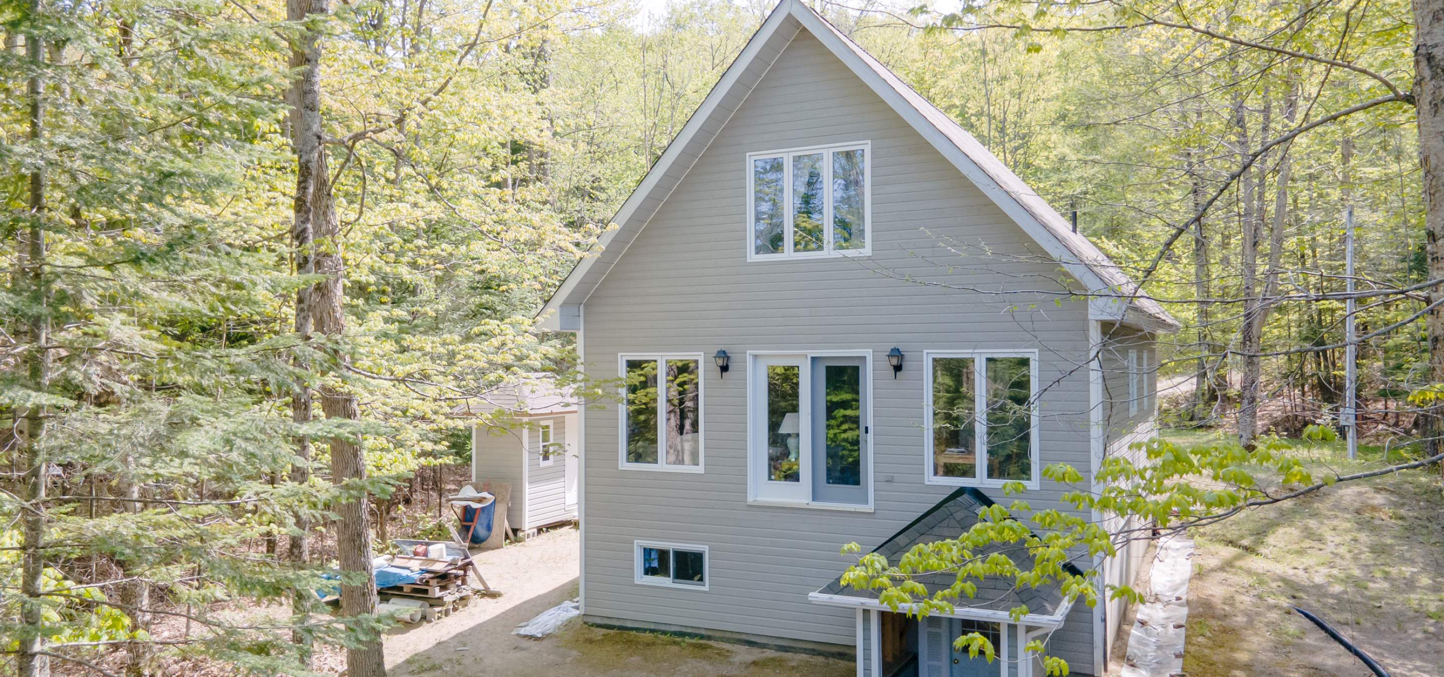 Grey with white trim storey and a half cottage on a level lot surrounded by springtime hardwood trees.
